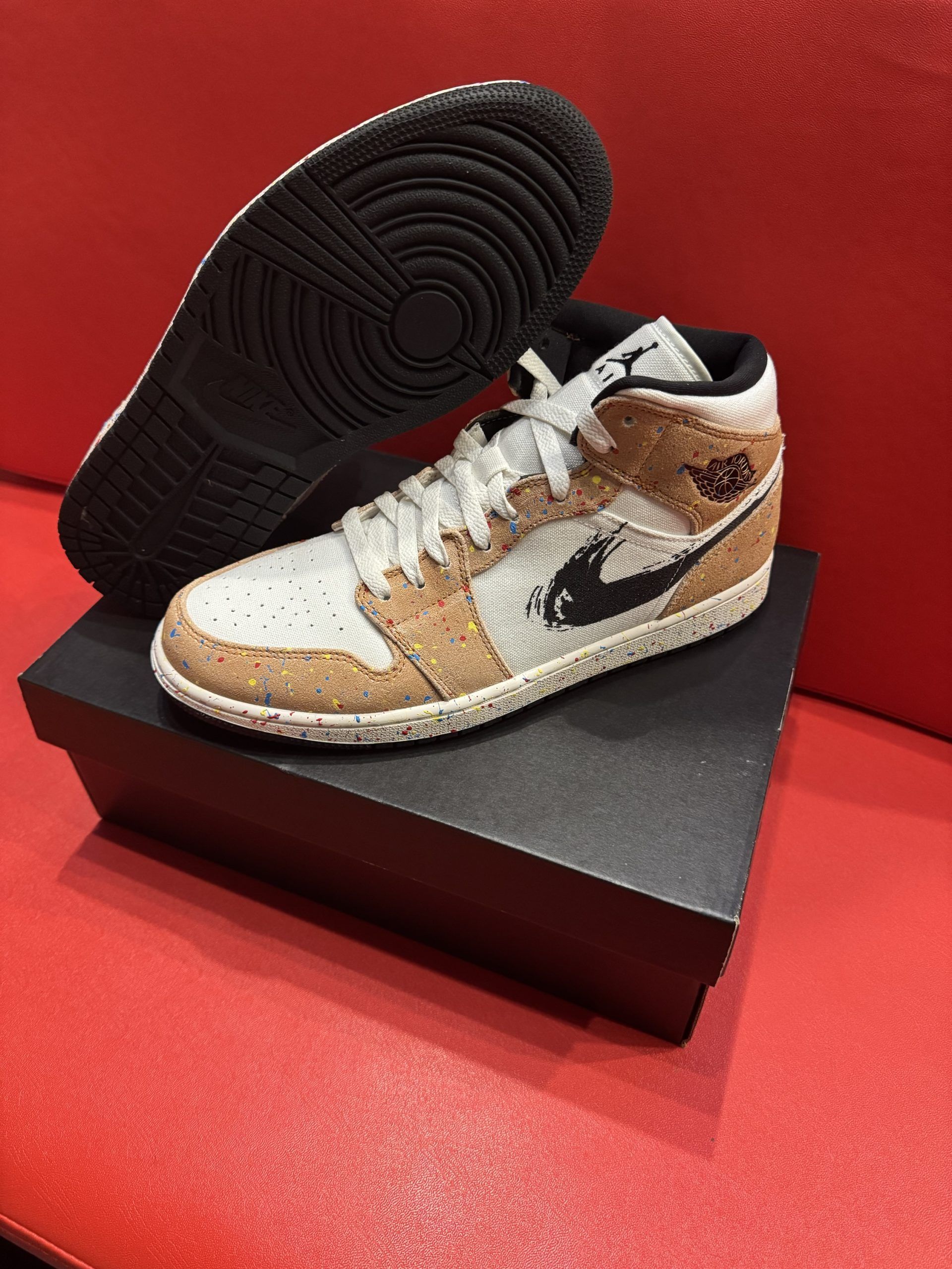 Air Jordan 1 Mid sneaker with cork-like panels, black swoosh, and black sole, resting on a black box.