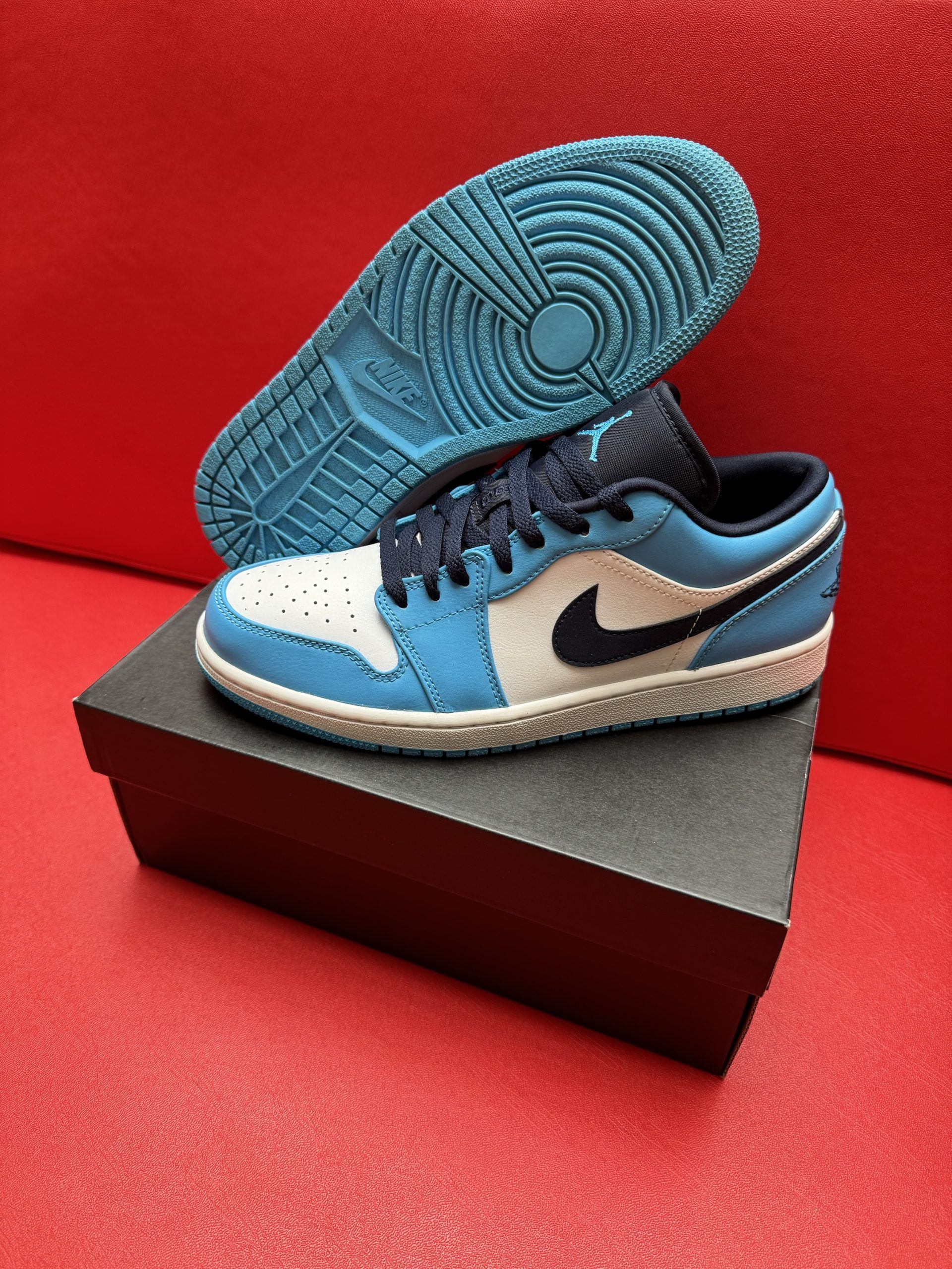 Air Jordan 1 low sneaker in blue and white on a black box, sole in background on red surface.