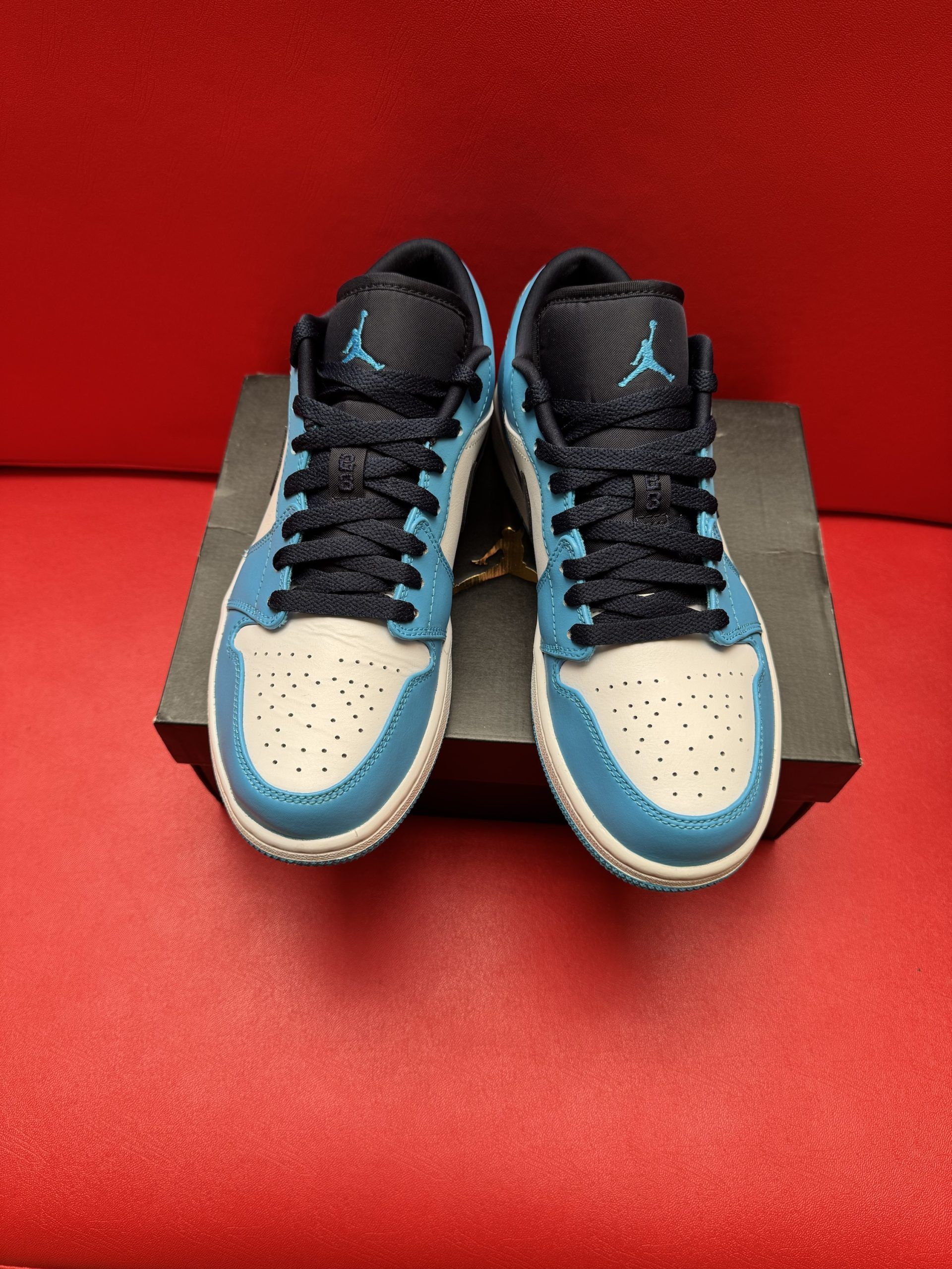 Air Jordan 1 low sneaker in blue and white on a black box, sole in background on red surface.