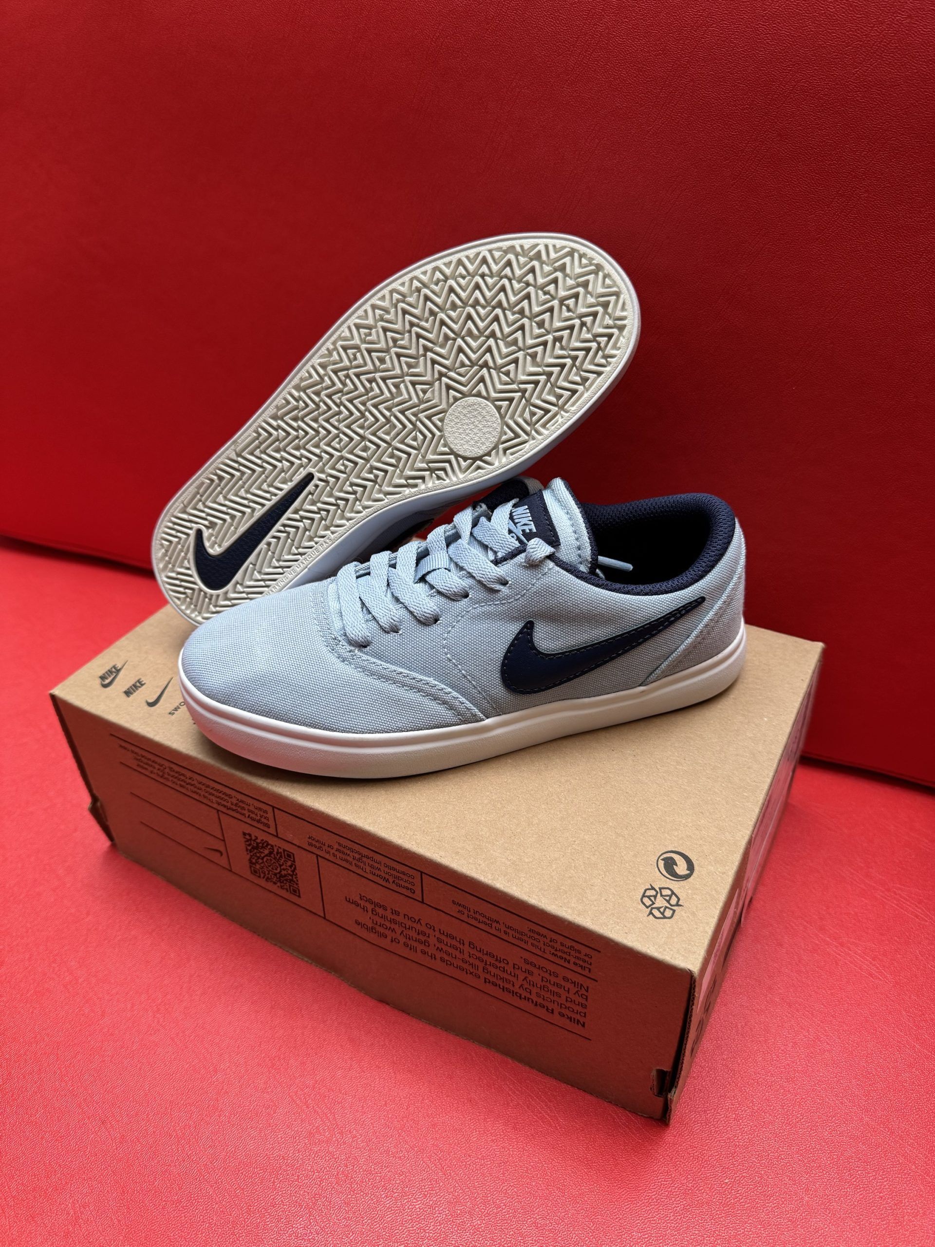 Light blue Nike sneakers with dark blue swoosh on a brown cardboard box, red background.