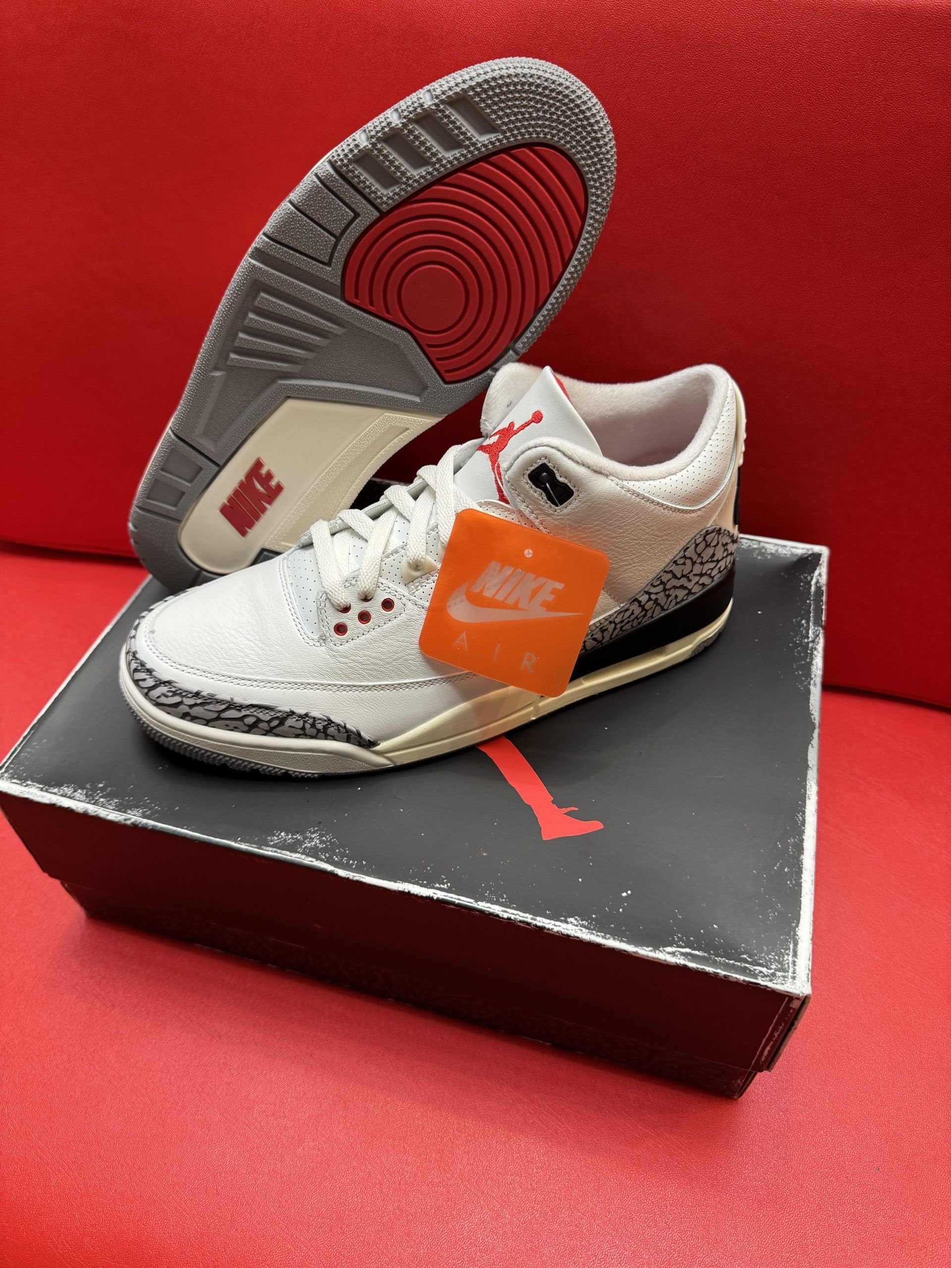 White and gray Air Jordan 3 sneakers with red accents and a box on a red surface.