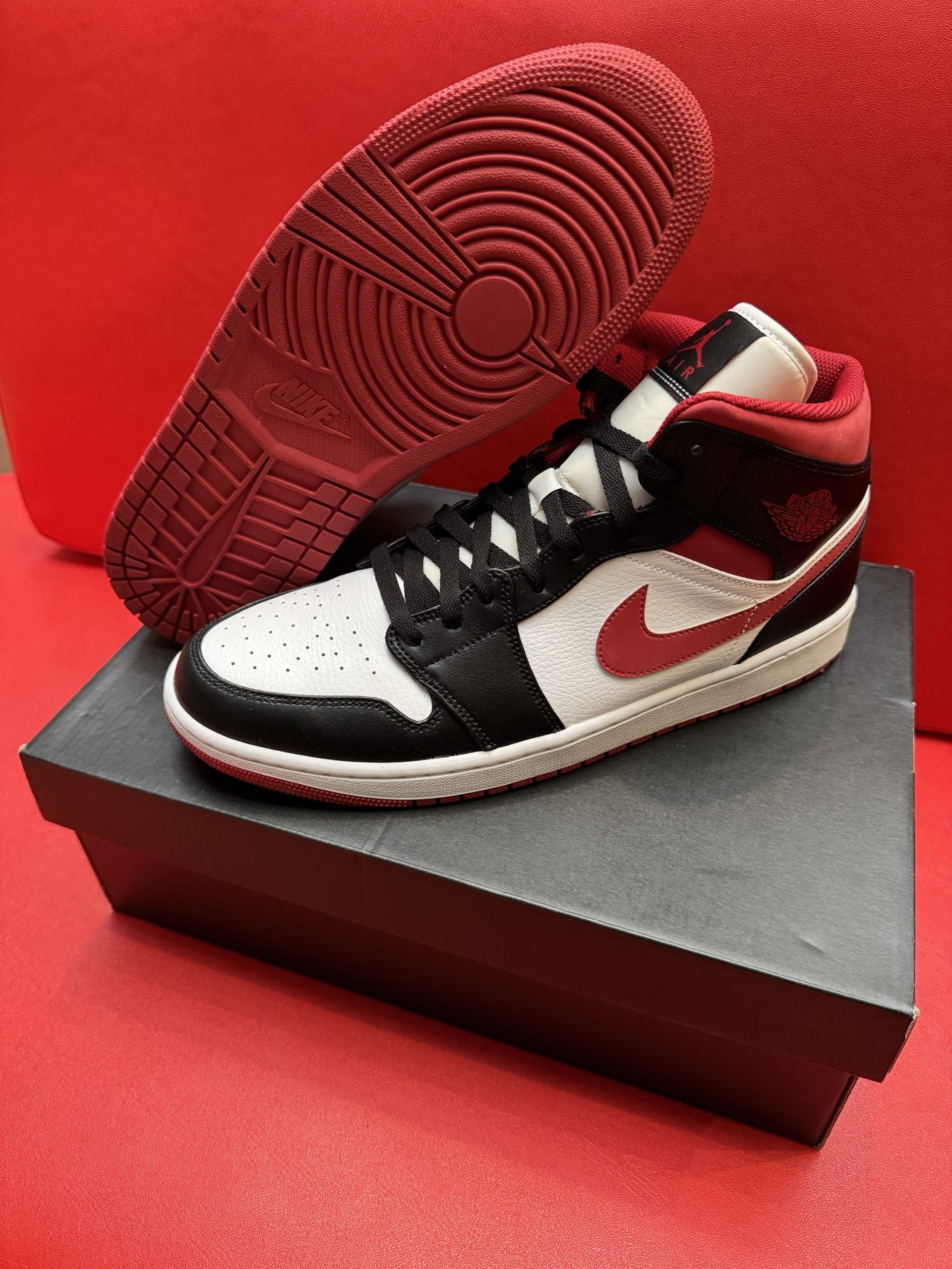 Air Jordan 1 Mid sneaker in black, red, and white on a black box, with the sole visible. Red background.