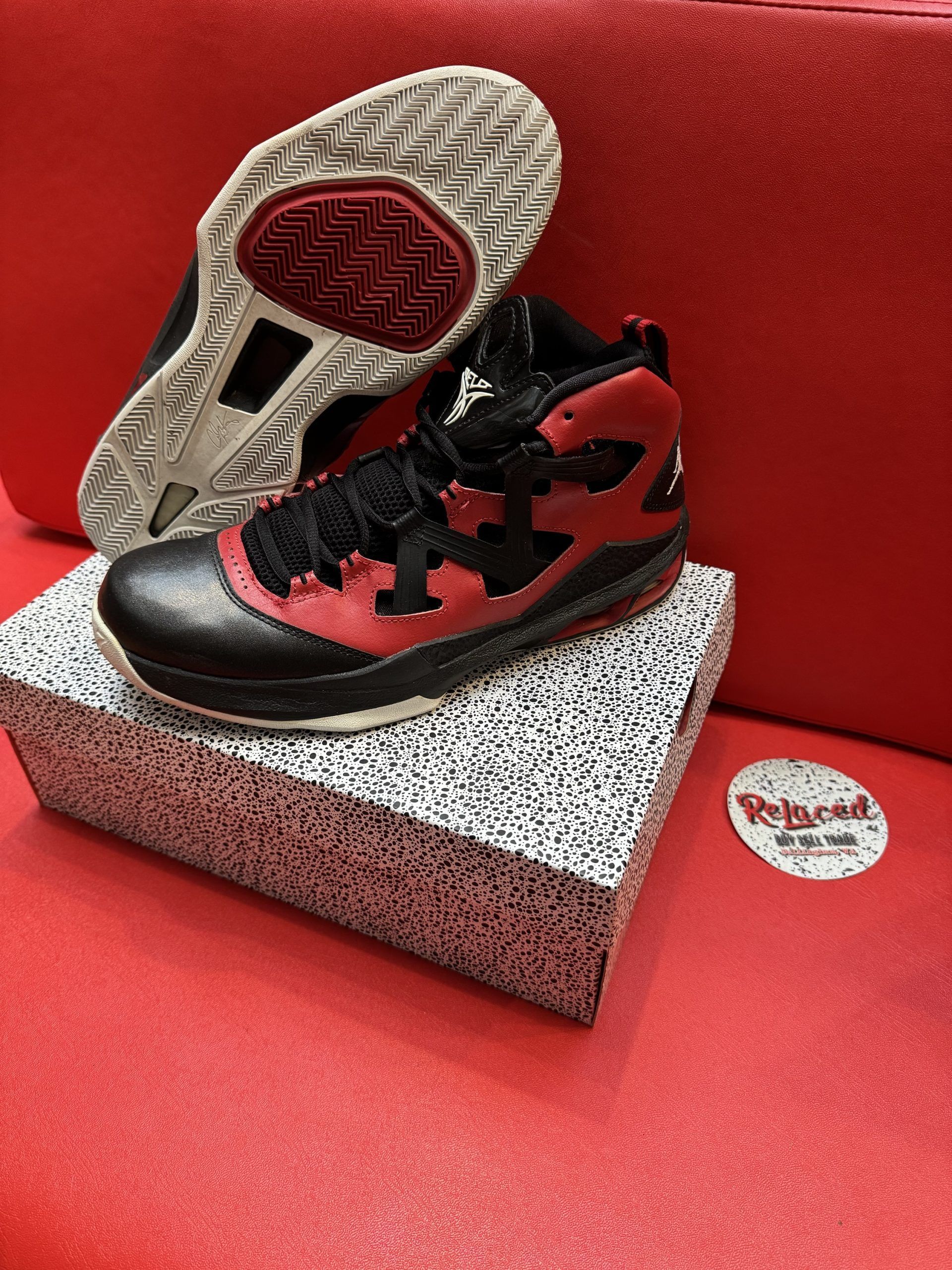 Black and red basketball shoe, top view, sitting on speckled box, red backdrop. Underside of shoe visible.