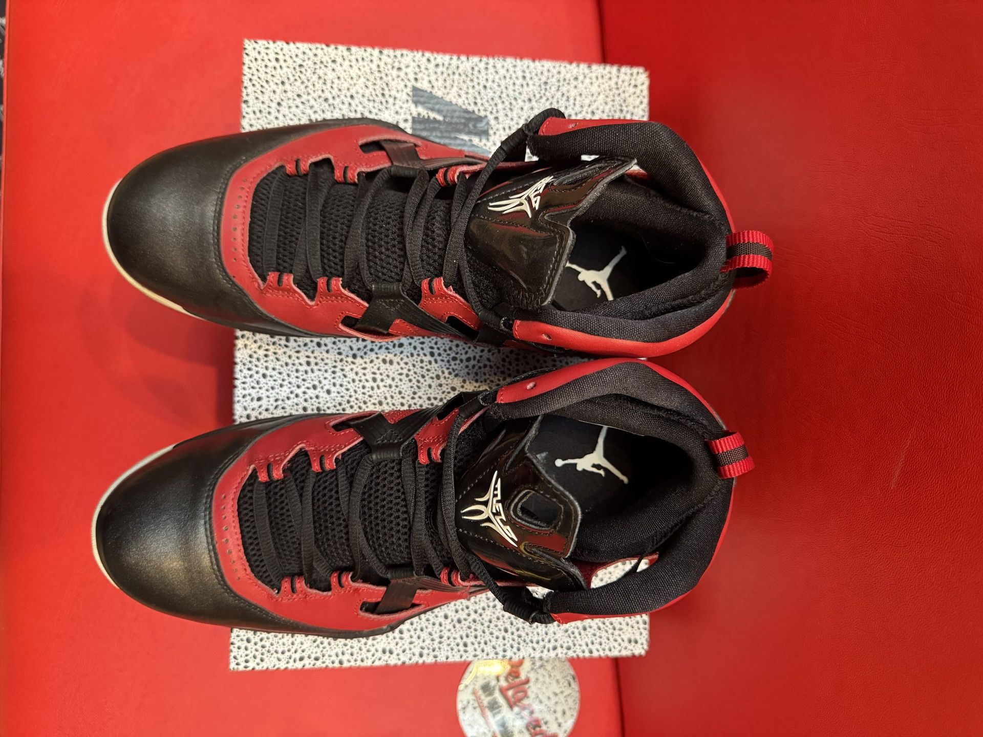 Black and red basketball shoe, top view, sitting on speckled box, red backdrop. Underside of shoe visible.