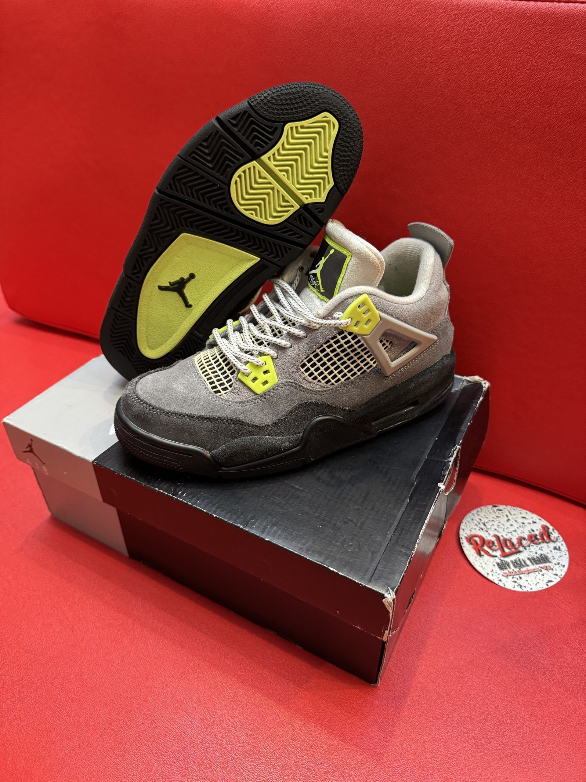 Gray Air Jordan 4 sneakers with black sole and yellow accents, on a box and red backdrop.