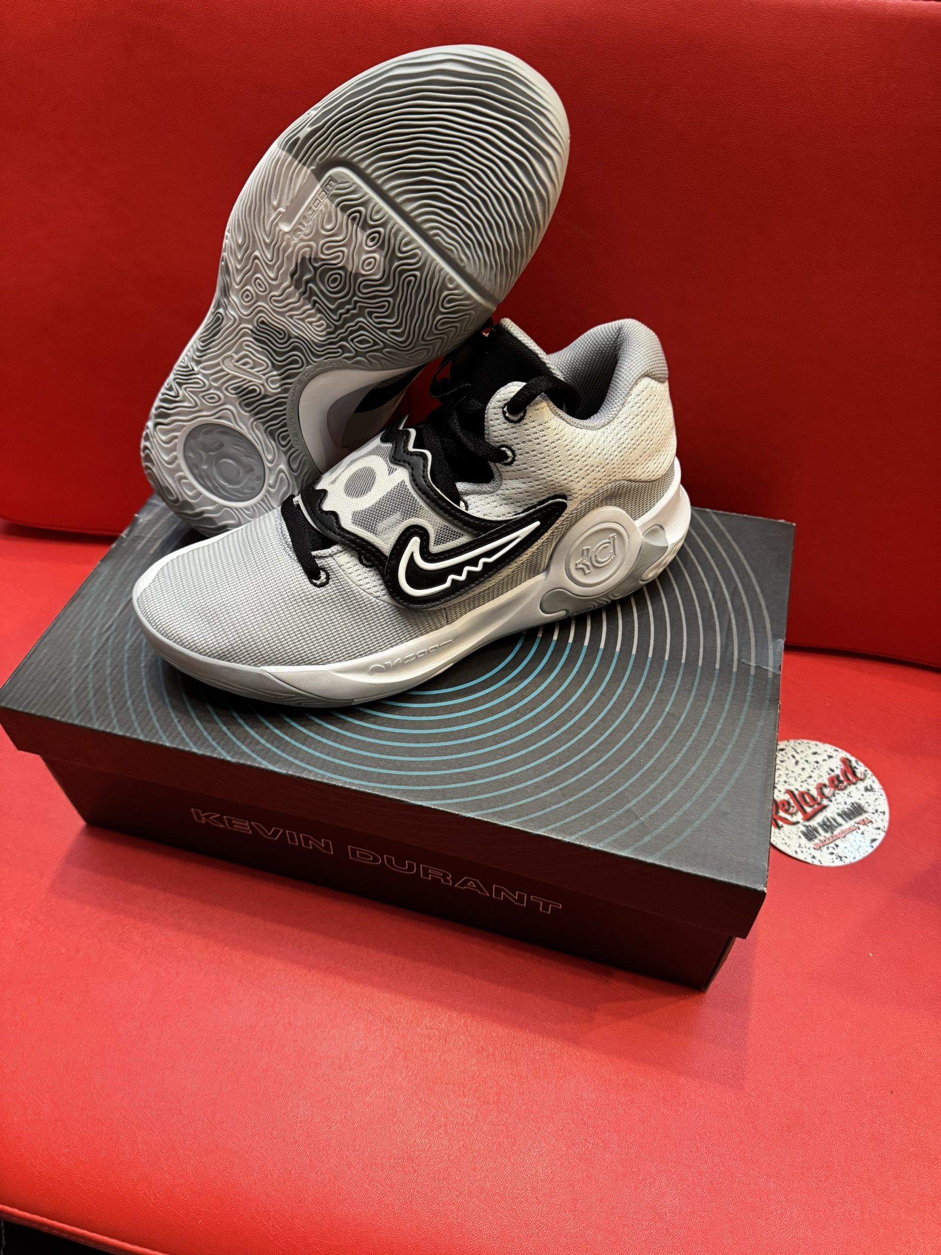 Basketball shoe on top of a shoe box. The shoe is white with black accents and a black swoosh. The sole is visible.