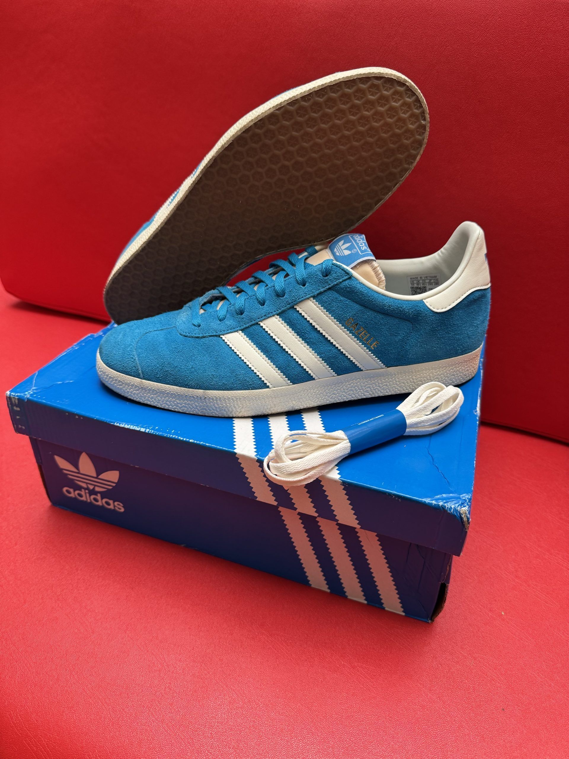 Blue Adidas Gazelle sneaker on box with extra laces and insole on red surface.