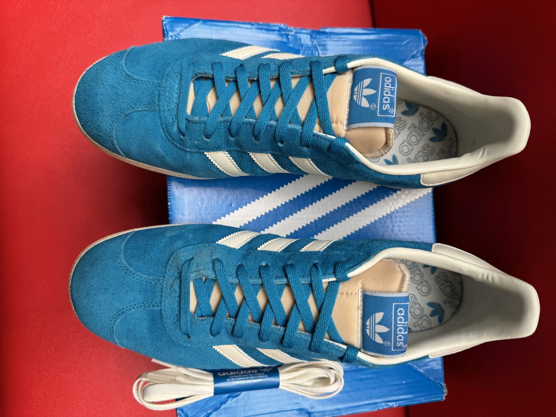 Blue Adidas Gazelle sneaker on box with extra laces and insole on red surface.