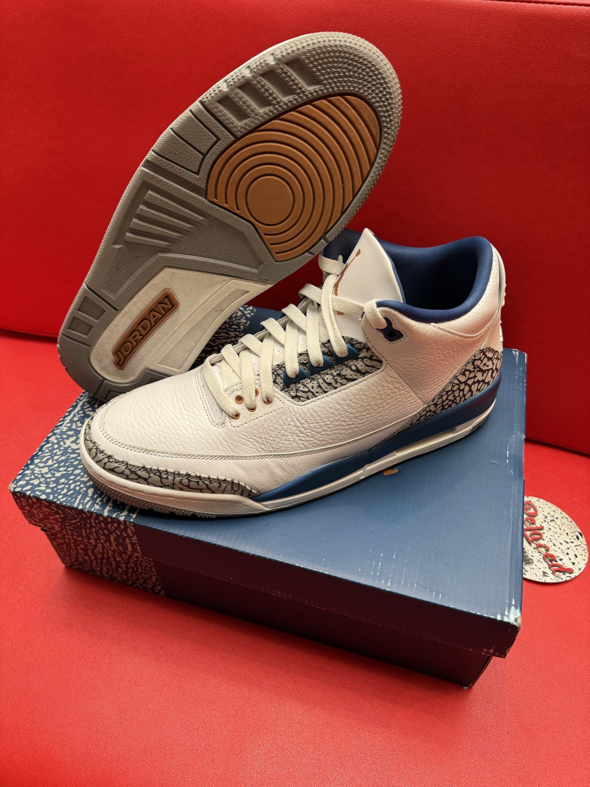 Pair of white and blue Air Jordan 3 sneakers with grey bottoms on a blue box, set on a red surface.