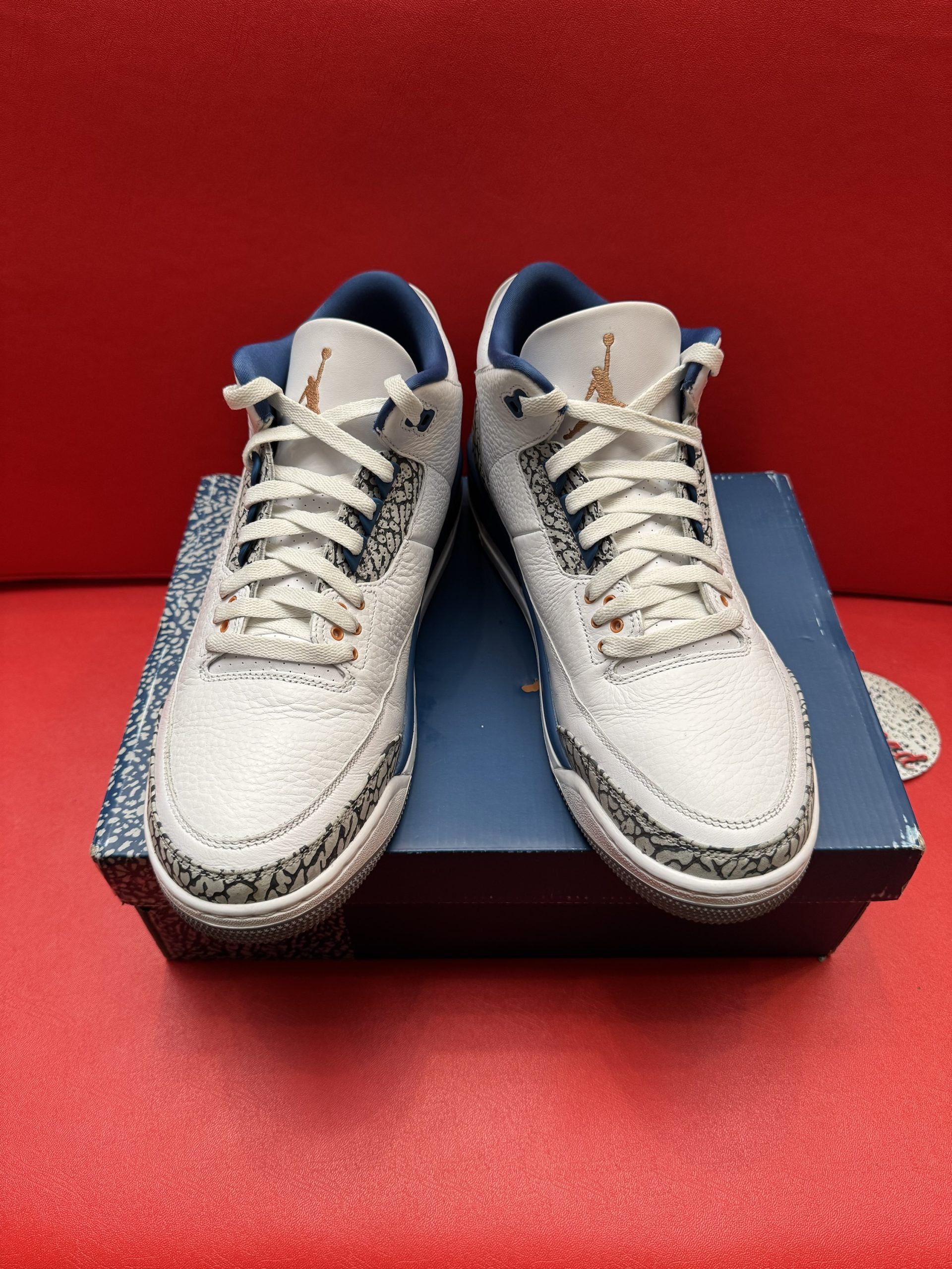 Pair of white and blue Air Jordan 3 sneakers with grey bottoms on a blue box, set on a red surface.