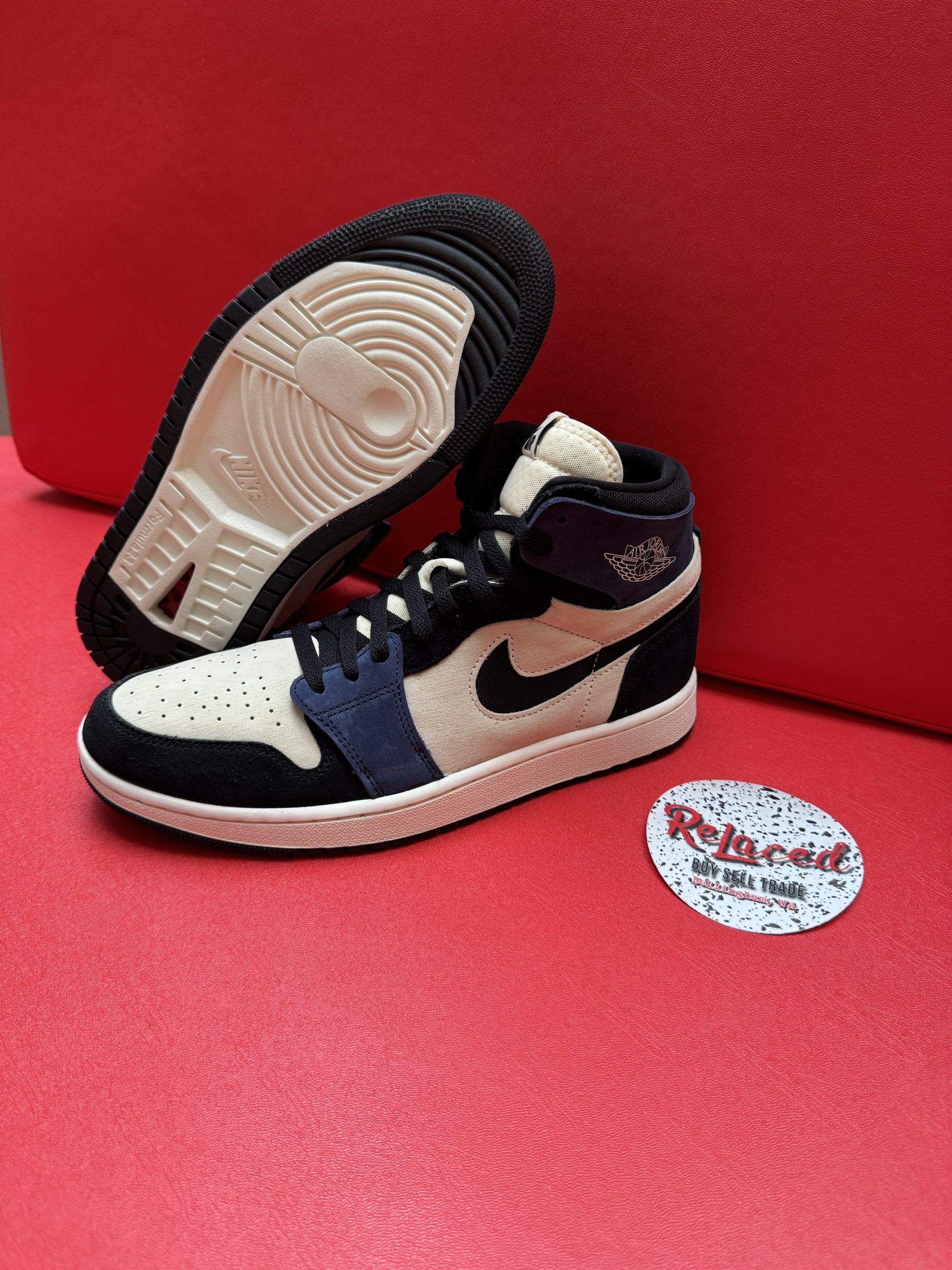 Air Jordan 1 sneakers: beige, navy, and black with a white sole, displayed on a red surface.
