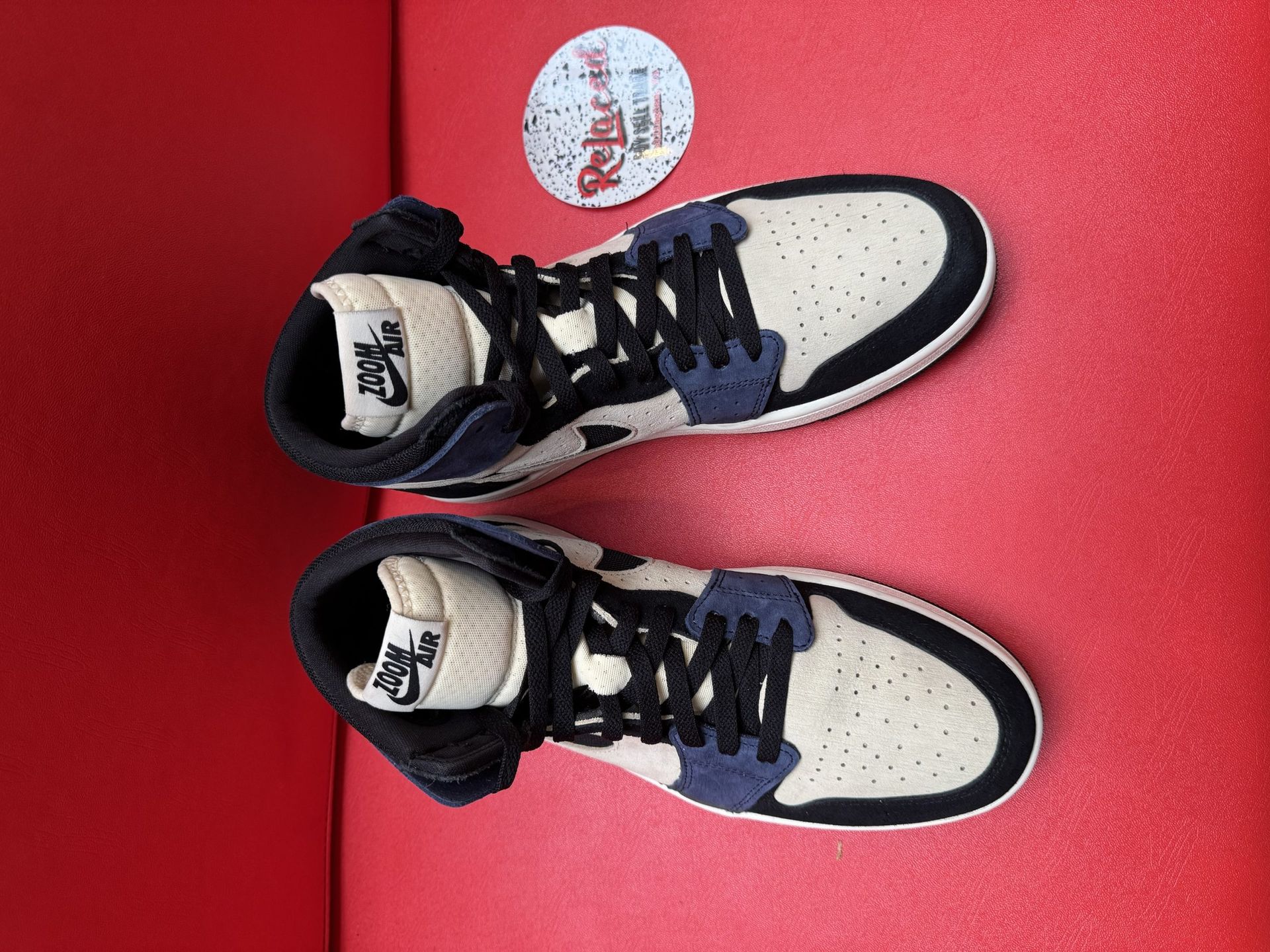 Air Jordan 1 sneakers: beige, navy, and black with a white sole, displayed on a red surface.