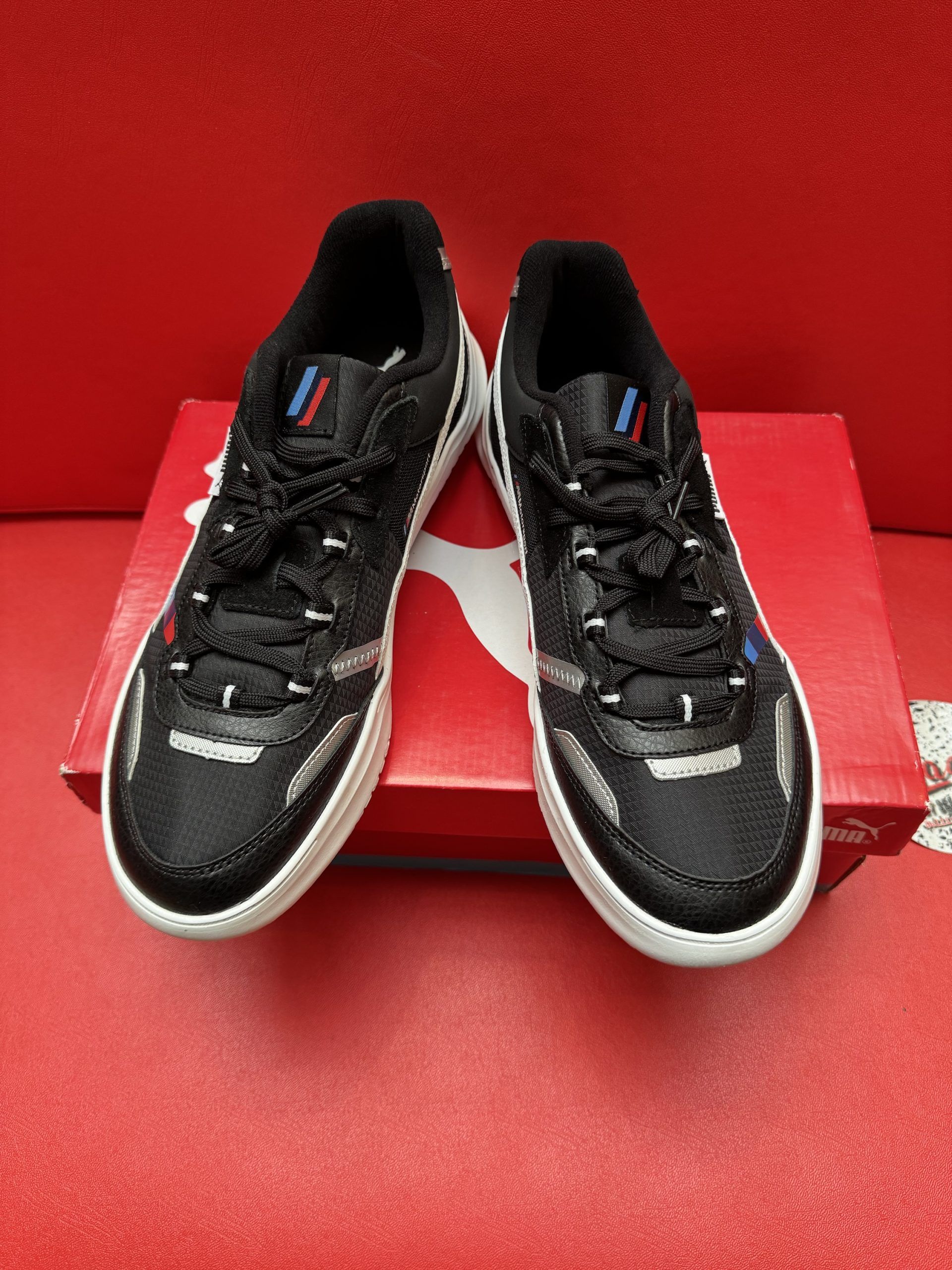 Black Puma shoe with white stripe, on red box and background. The sole is visible.