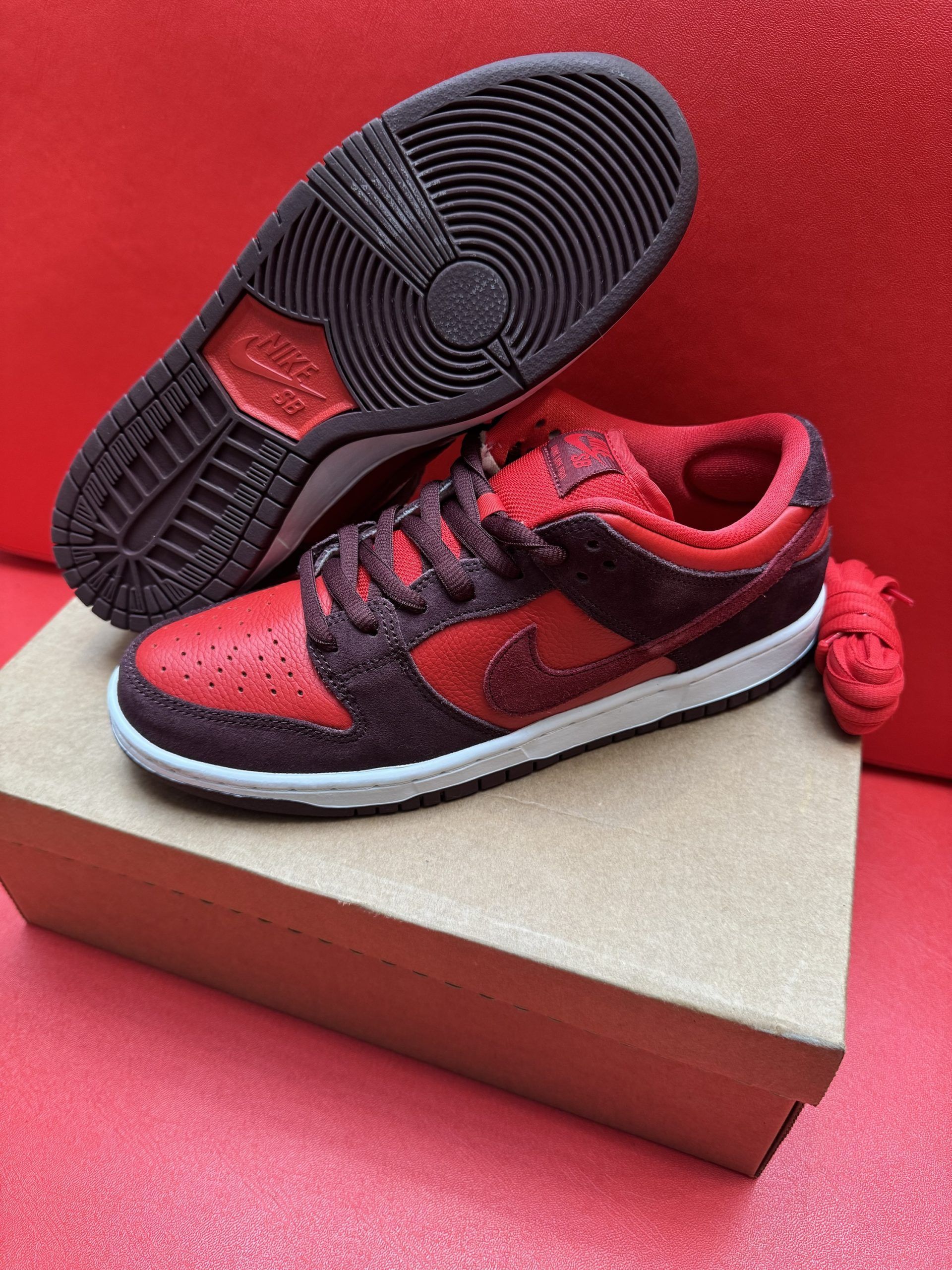 Red and black Nike Dunk Low sneakers with white sole, on a cardboard box against a red background.