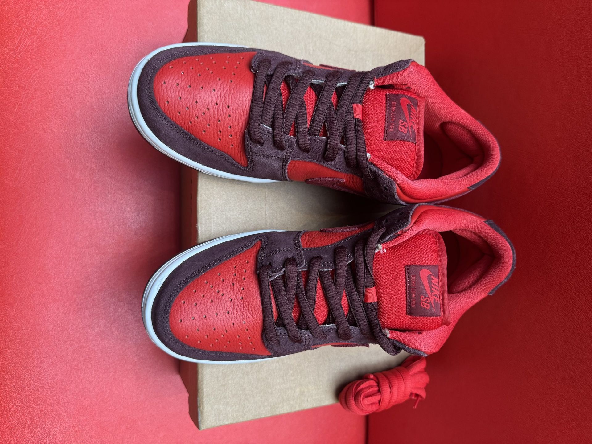 Red and black Nike Dunk Low sneakers with white sole, on a cardboard box against a red background.