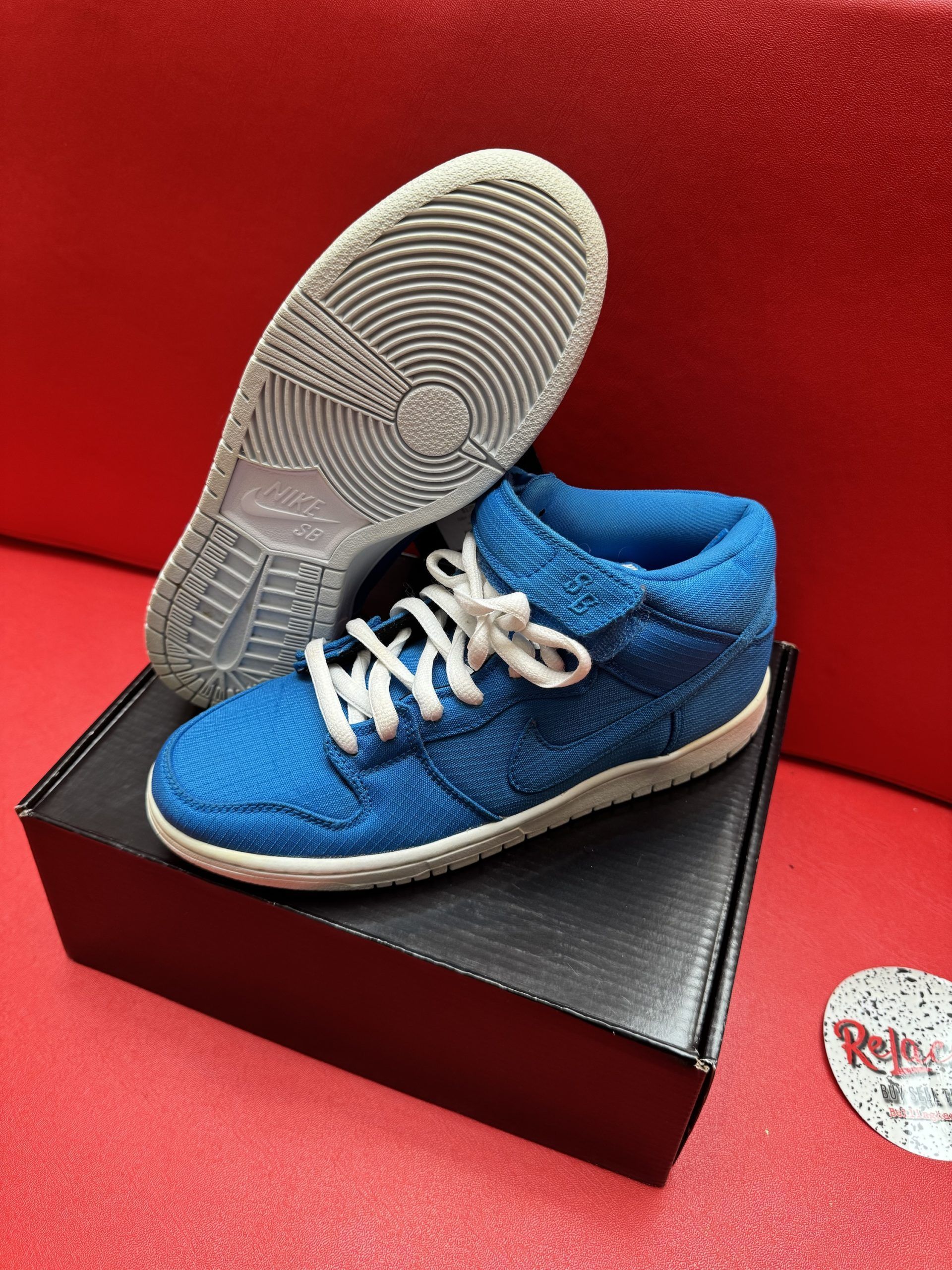 Blue Nike Dunk high-top sneaker with white laces, resting on a black shoebox, against a red background.