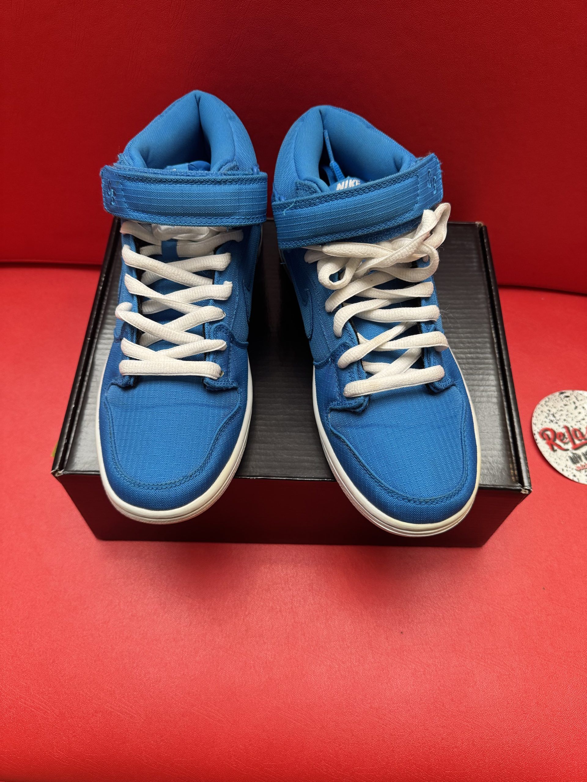 Blue Nike Dunk high-top sneaker with white laces, resting on a black shoebox, against a red background.