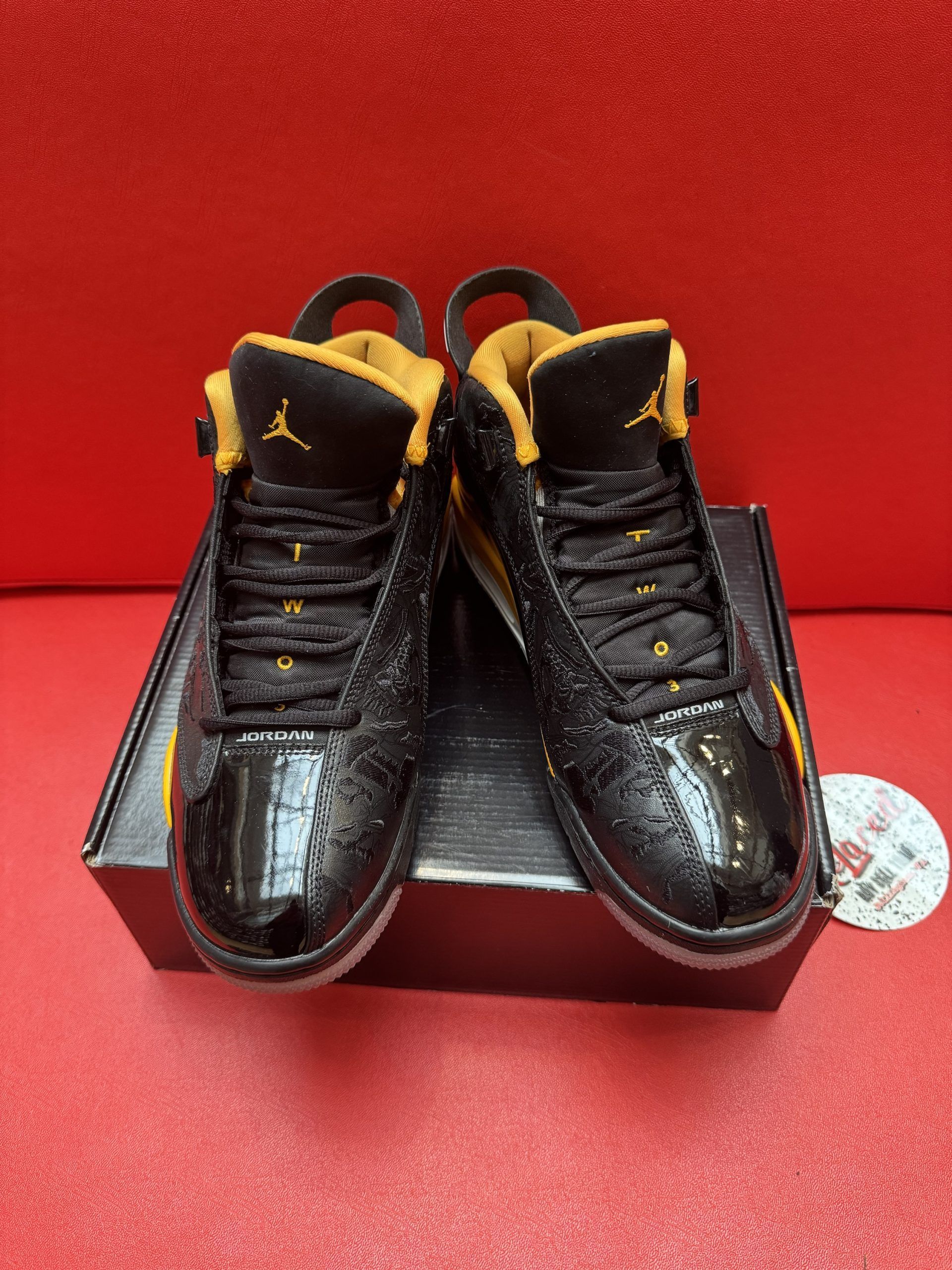 Black and yellow Air Jordan sneakers on a black box, with a red background and a logo sticker.