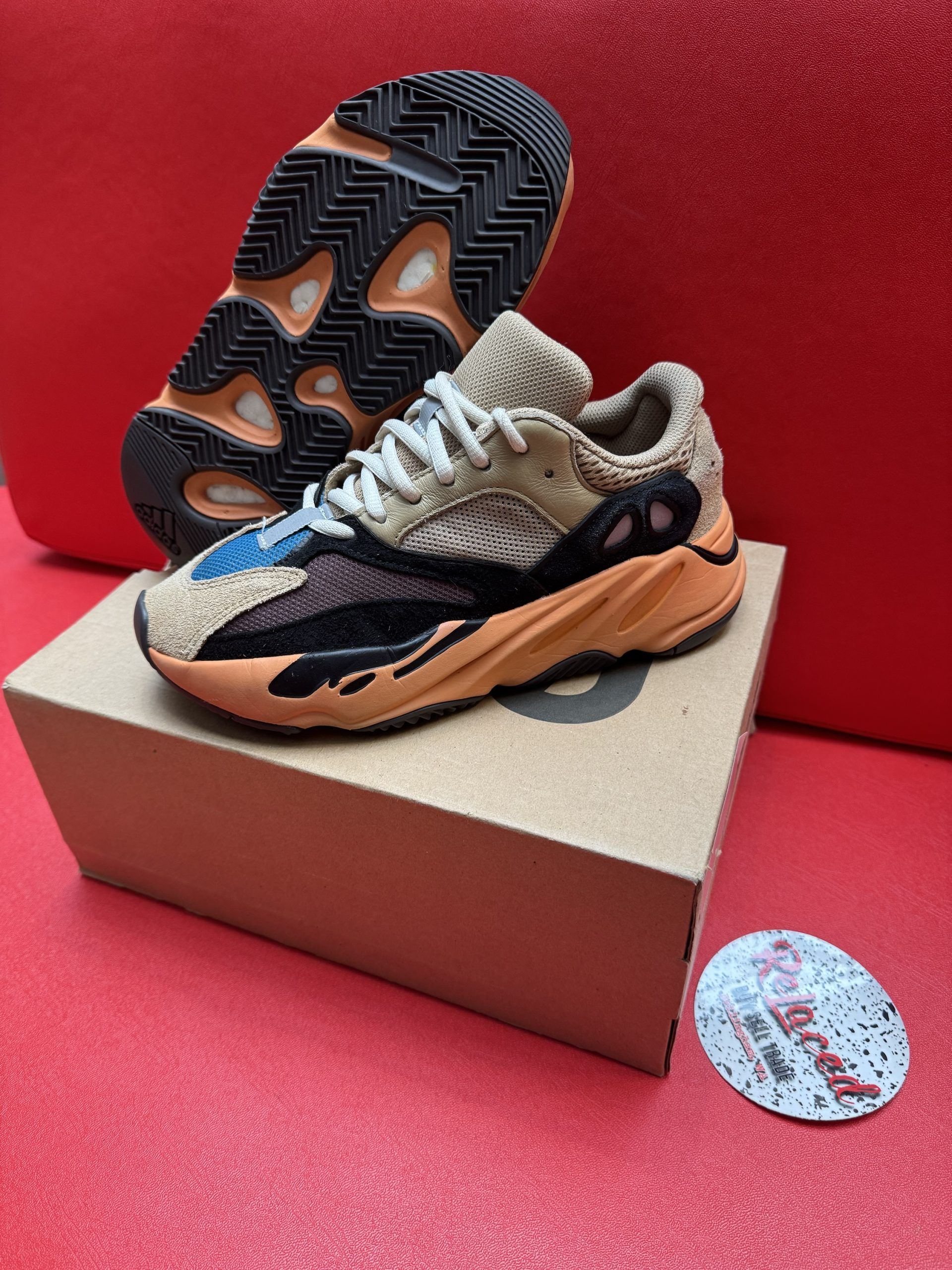 Yeezy 700 sneakers in orange, black, tan, and blue on a box, sole up behind the shoe, on a red surface.