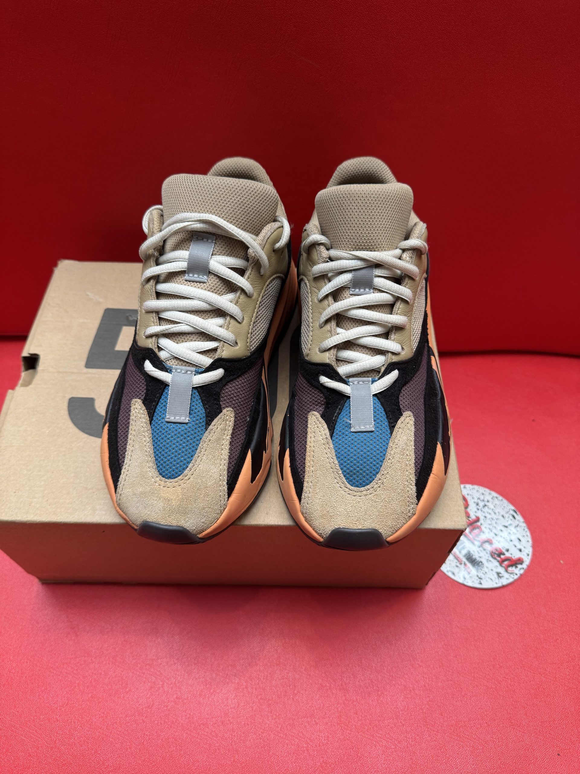 Yeezy 700 sneakers in orange, black, tan, and blue on a box, sole up behind the shoe, on a red surface.