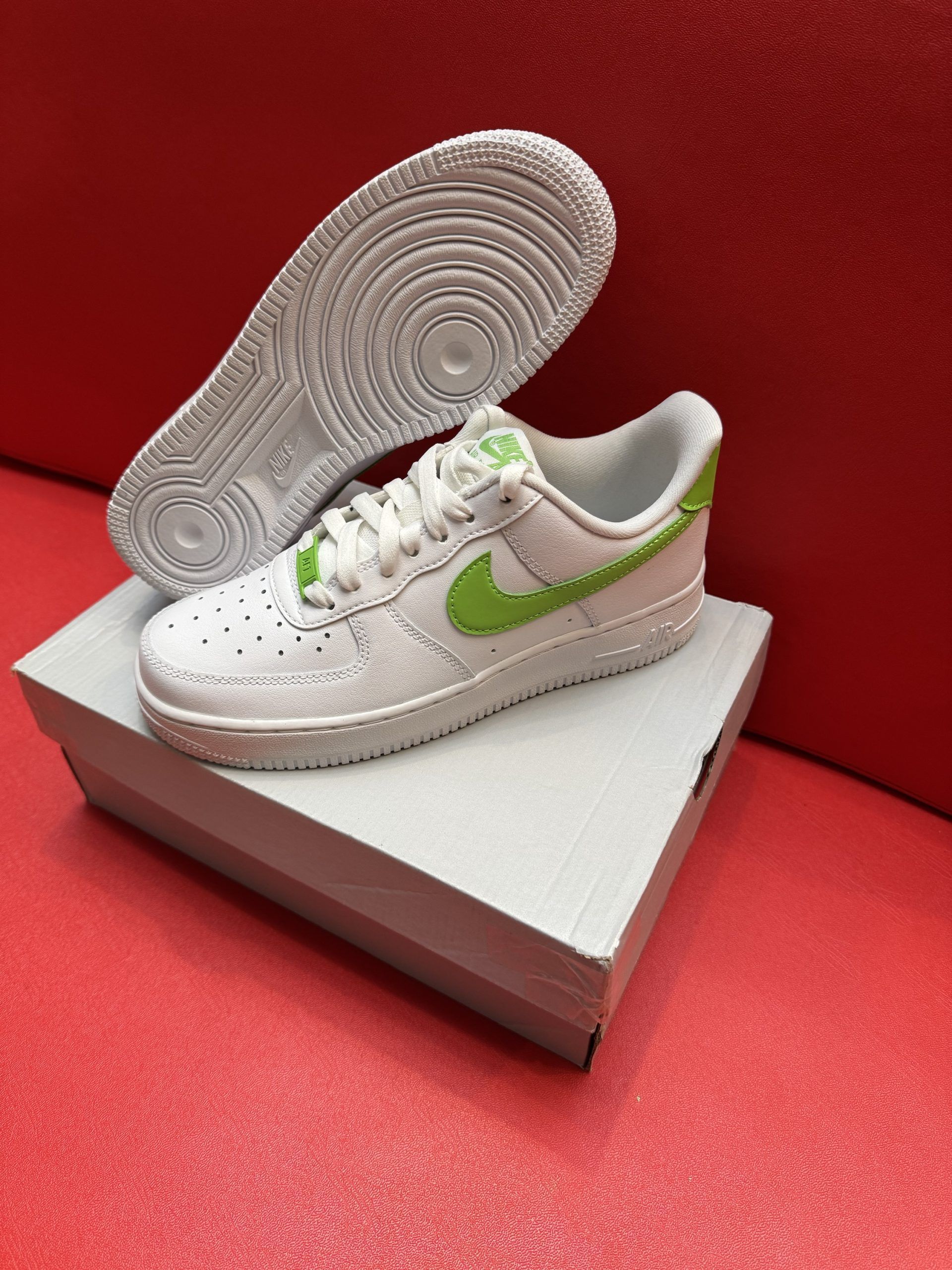 White Nike Air Force 1 sneakers with lime green accents, on top of a white box, and sole is displayed above on red surface.