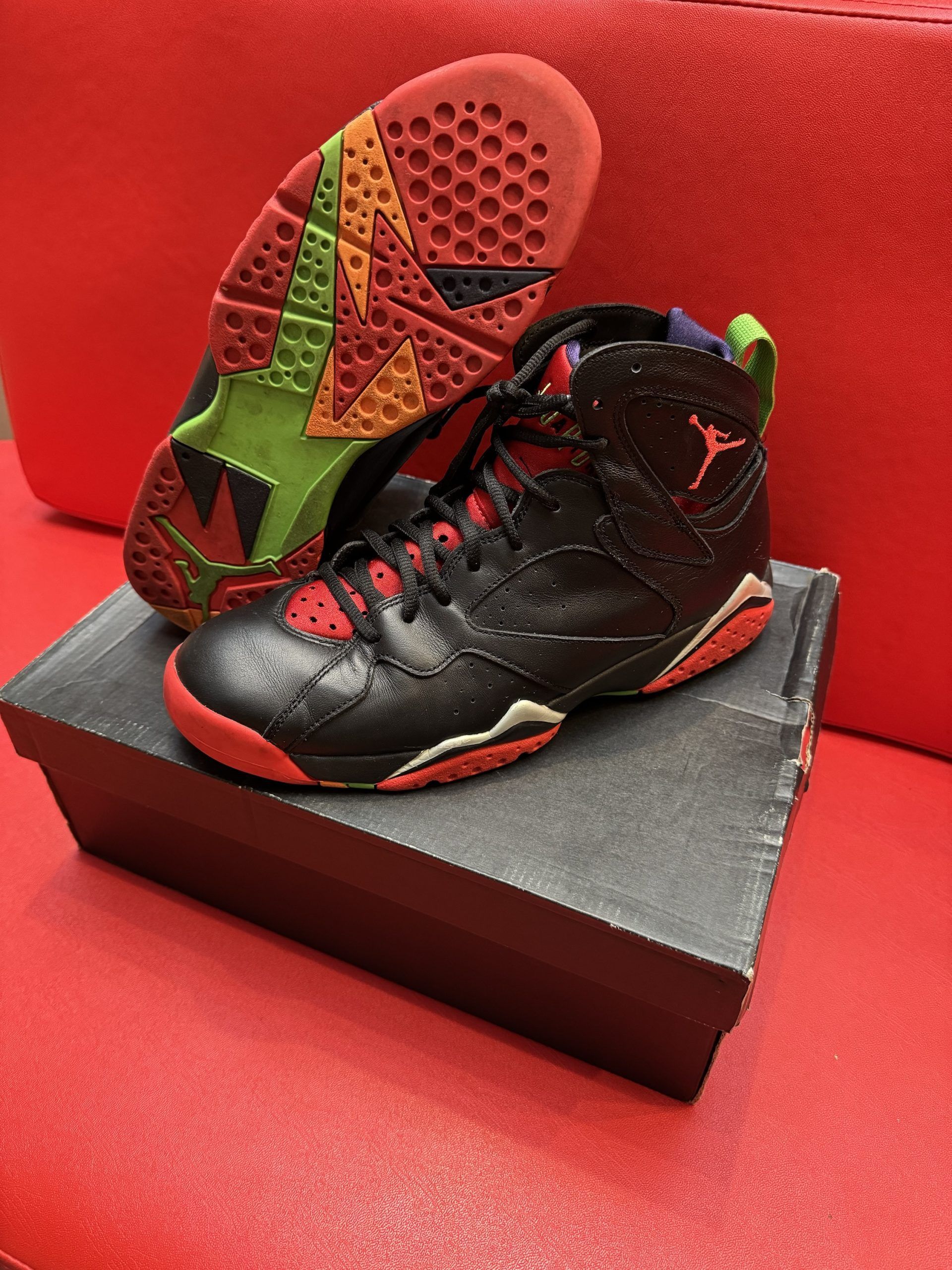Pair of black, red, and green Air Jordan 7 sneakers on a box, red background.
