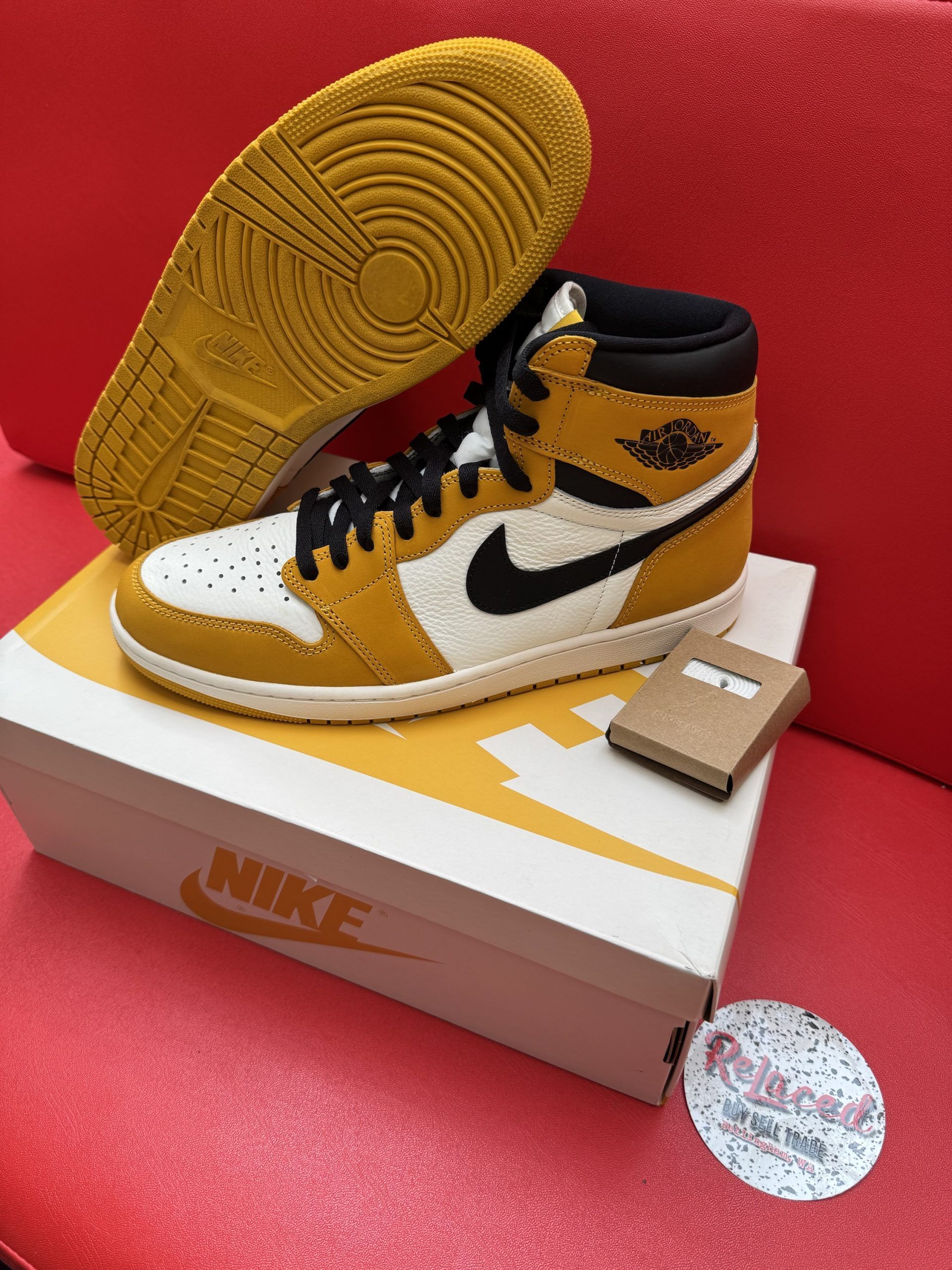 Yellow and white Air Jordan 1 sneaker on a box, with the sole visible, on a red surface.