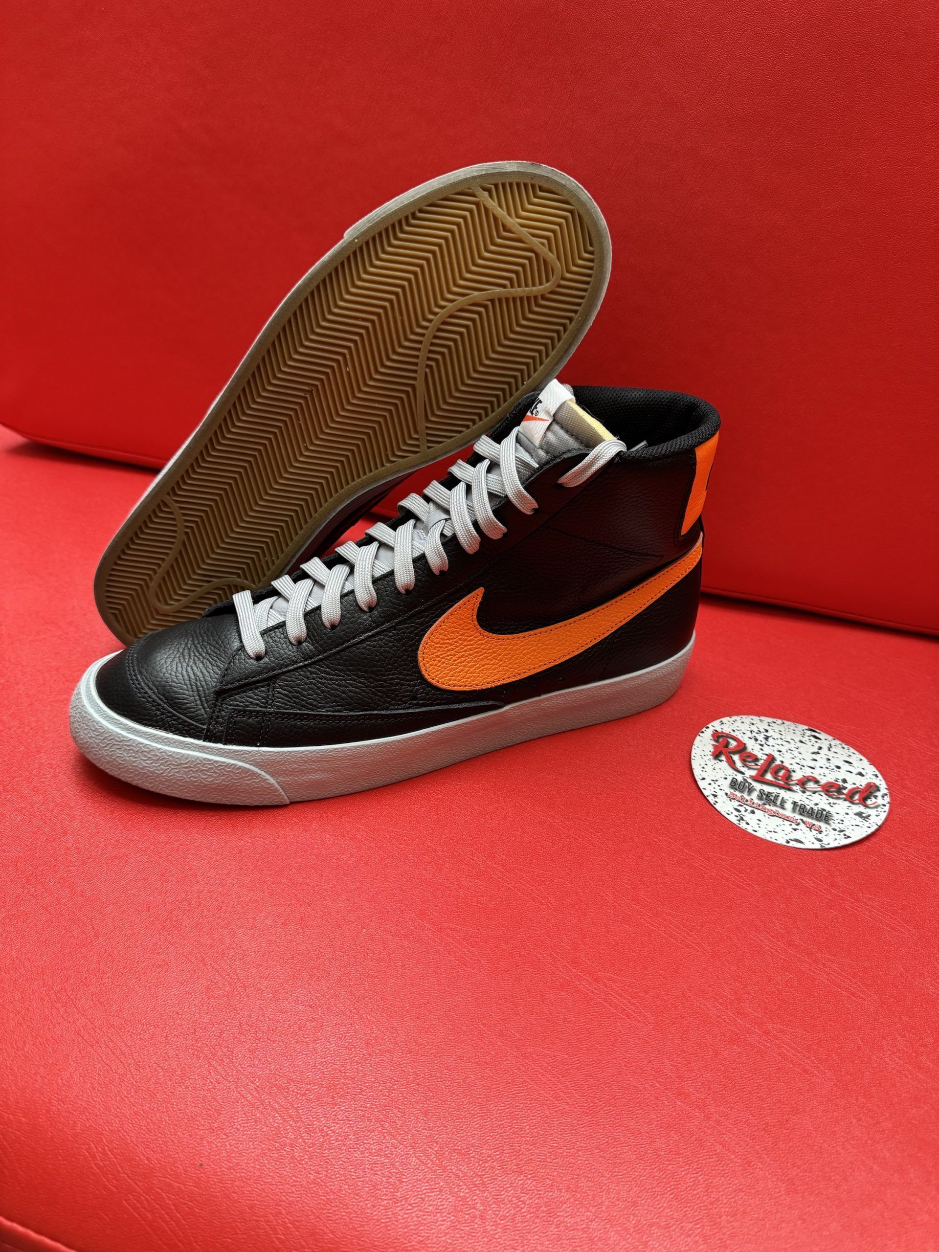 Black Nike Blazer sneaker with orange swoosh, silver laces, and off-white sole on a red surface.