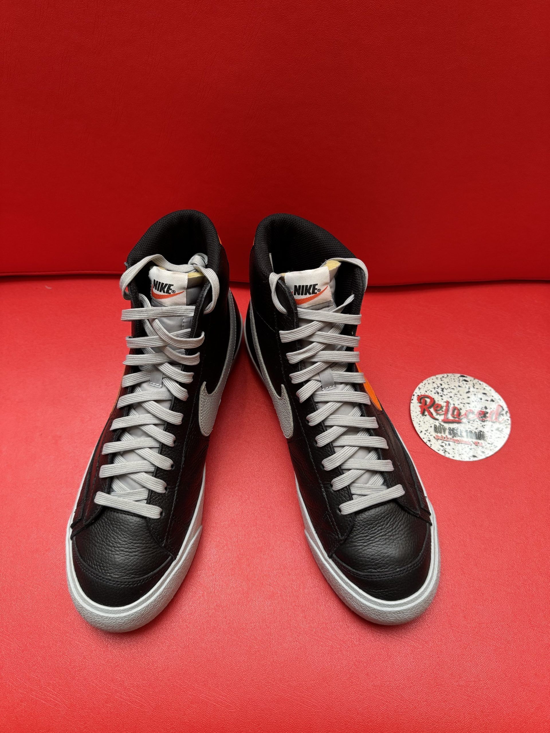 Black Nike Blazer sneaker with orange swoosh, silver laces, and off-white sole on a red surface.