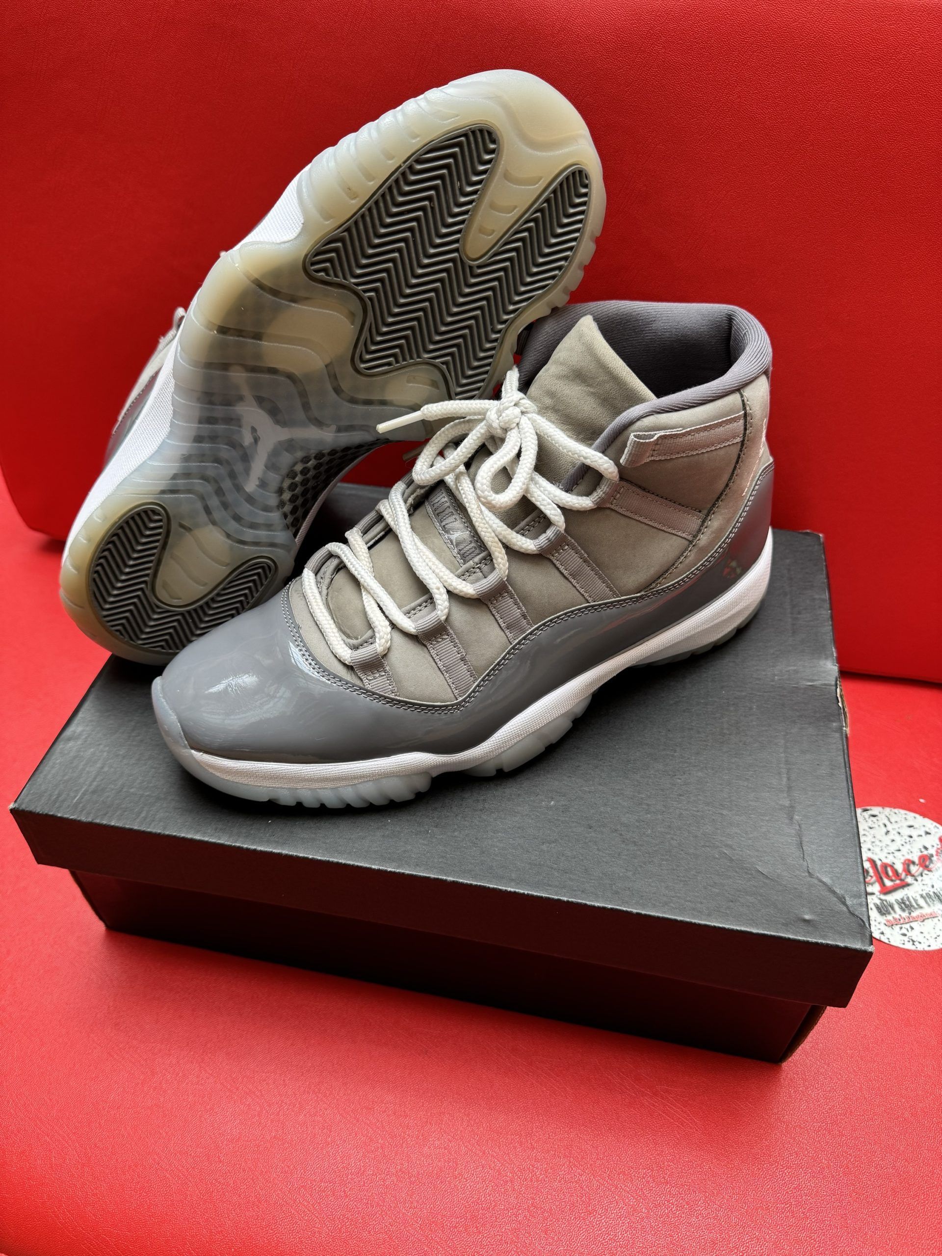 Pair of grey and white Air Jordan 11 Retro sneakers on a black shoebox, red background.