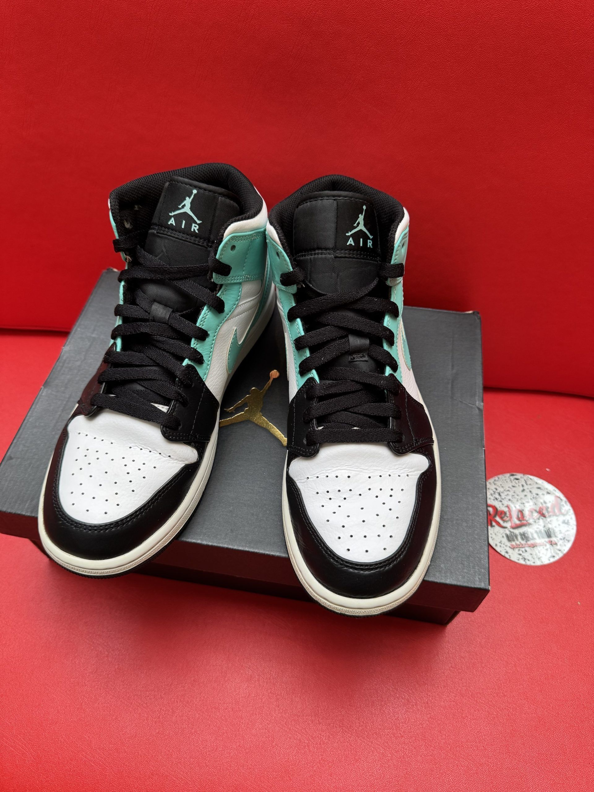 Air Jordan 1 Mid sneaker on black box, with a black and aqua color scheme and black sole, sitting on a red surface.