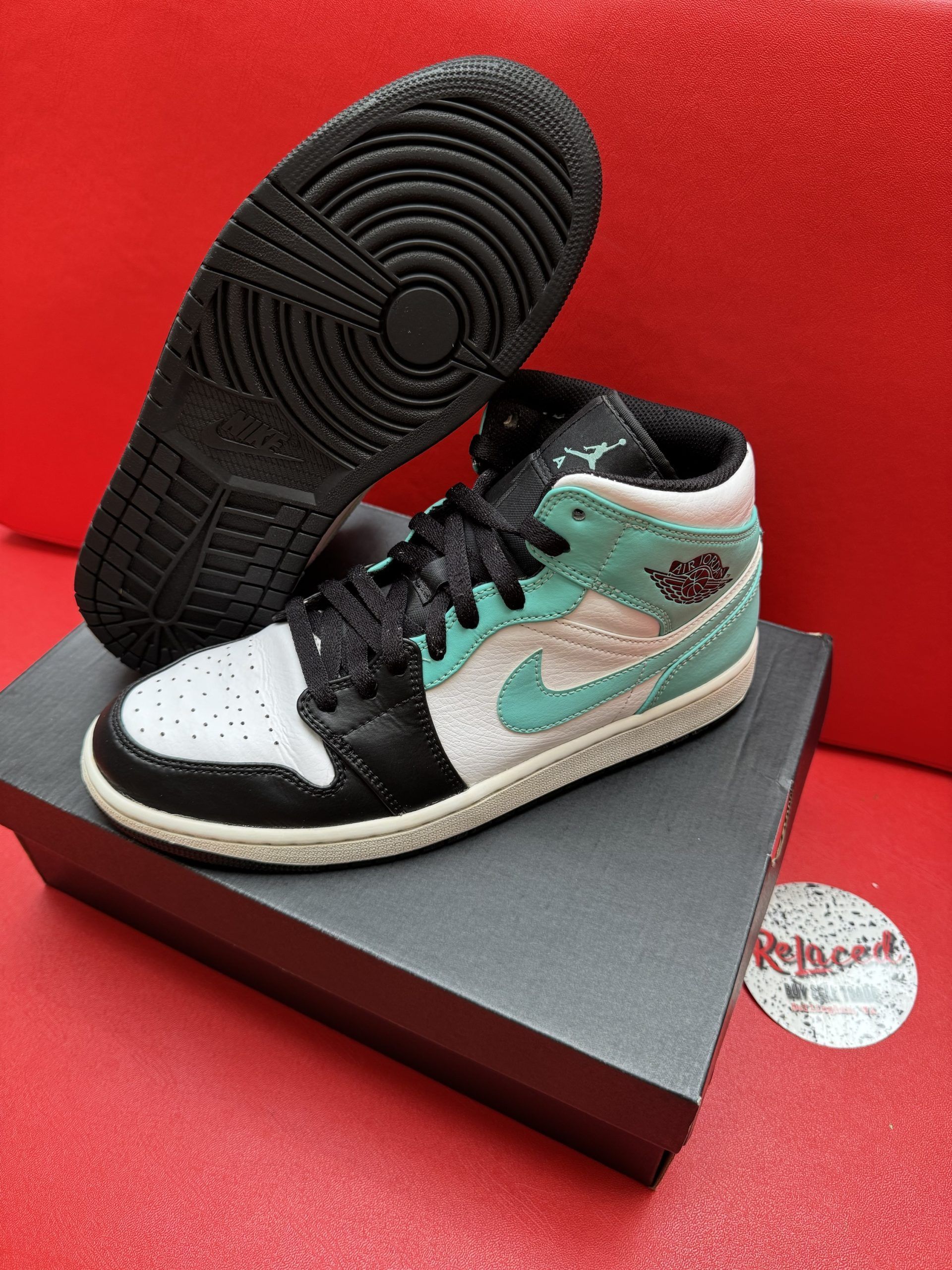 Air Jordan 1 Mid sneaker on black box, with a black and aqua color scheme and black sole, sitting on a red surface.