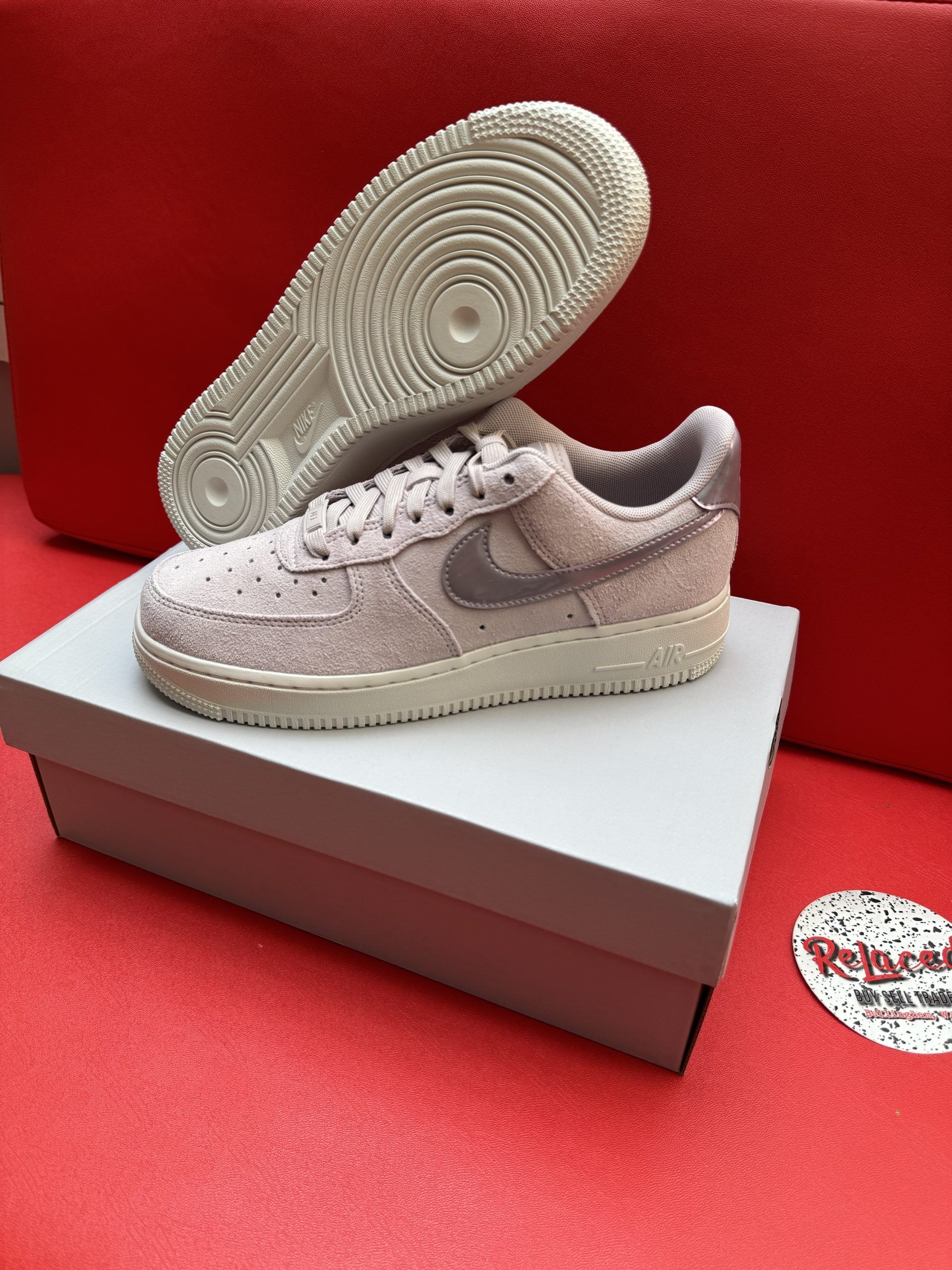 Nike Air Force 1 sneakers on a box, light pink, white sole, against red background.
