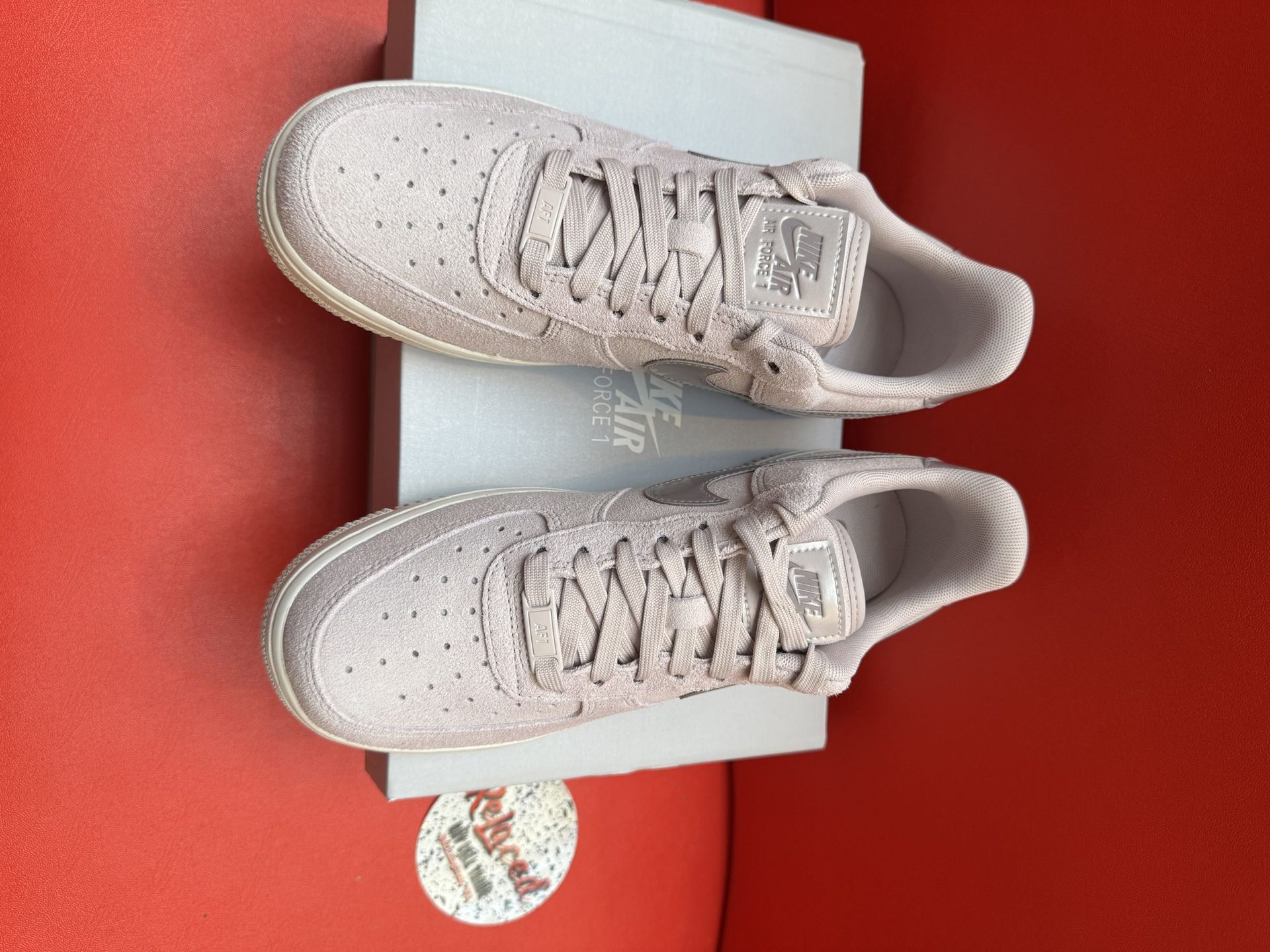 Nike Air Force 1 sneakers on a box, light pink, white sole, against red background.