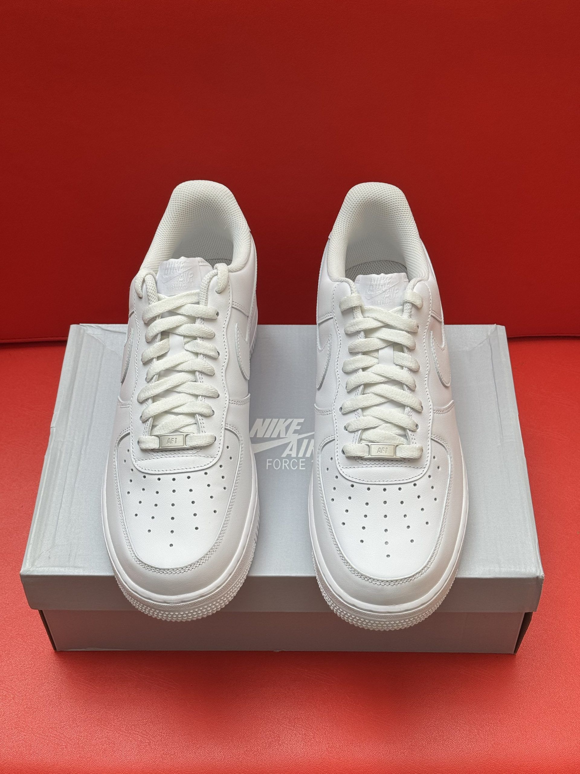White Nike Air Force 1 sneakers on a light gray box, resting on a red surface.