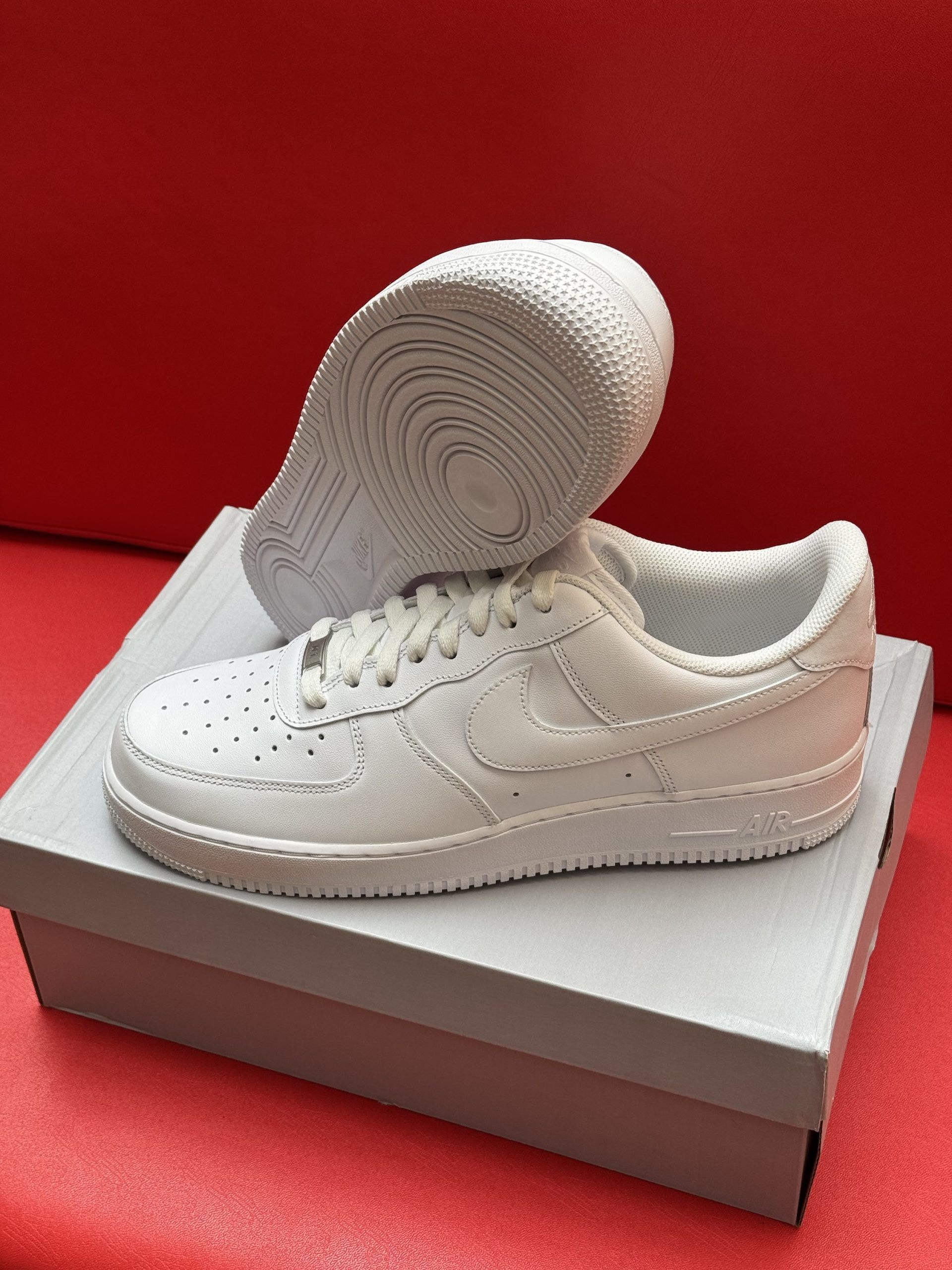 White Nike Air Force 1 sneakers on a light gray box, resting on a red surface.