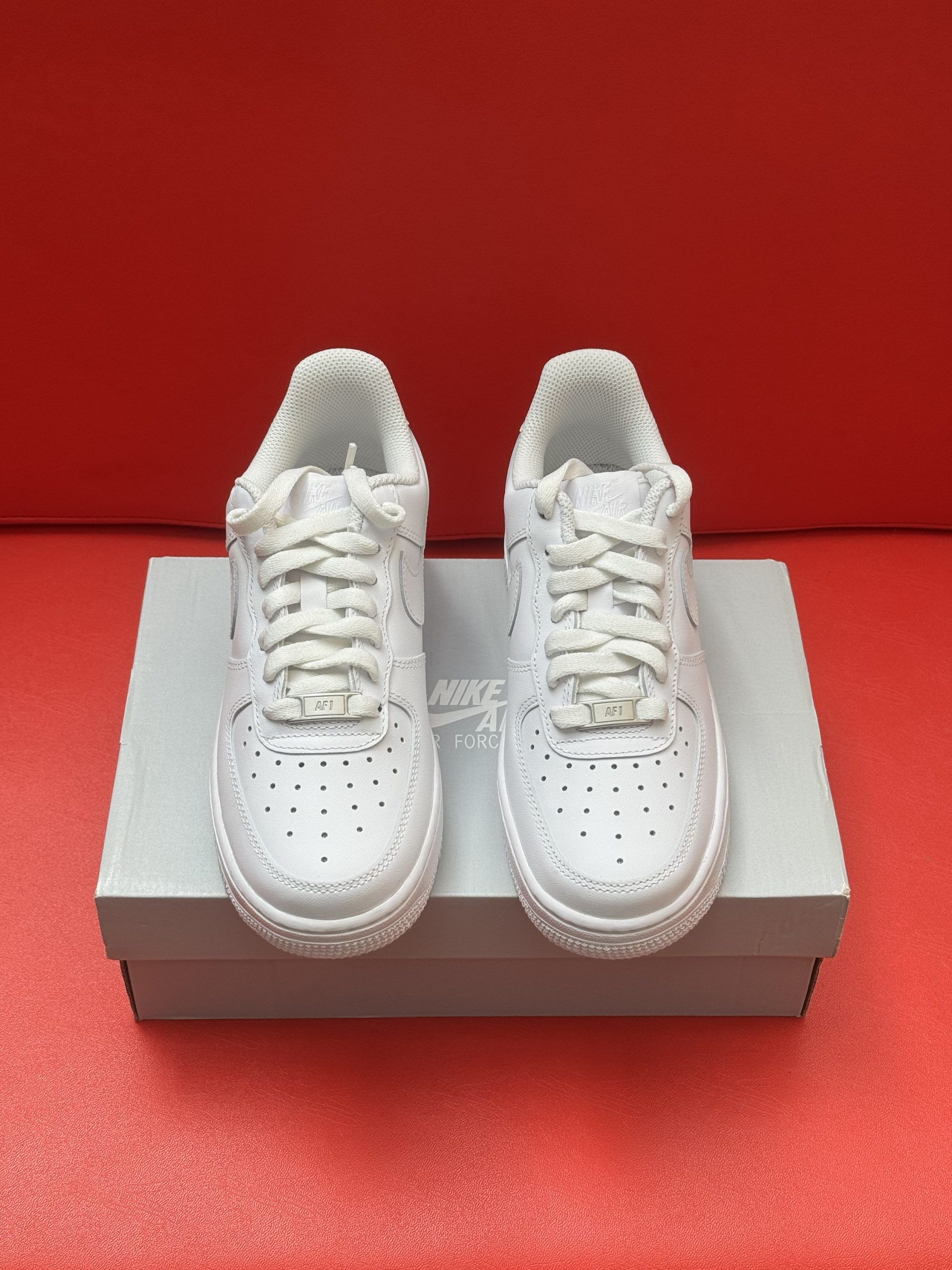 White Nike Air Force 1 sneakers on a box, one sole up, against a red backdrop.
