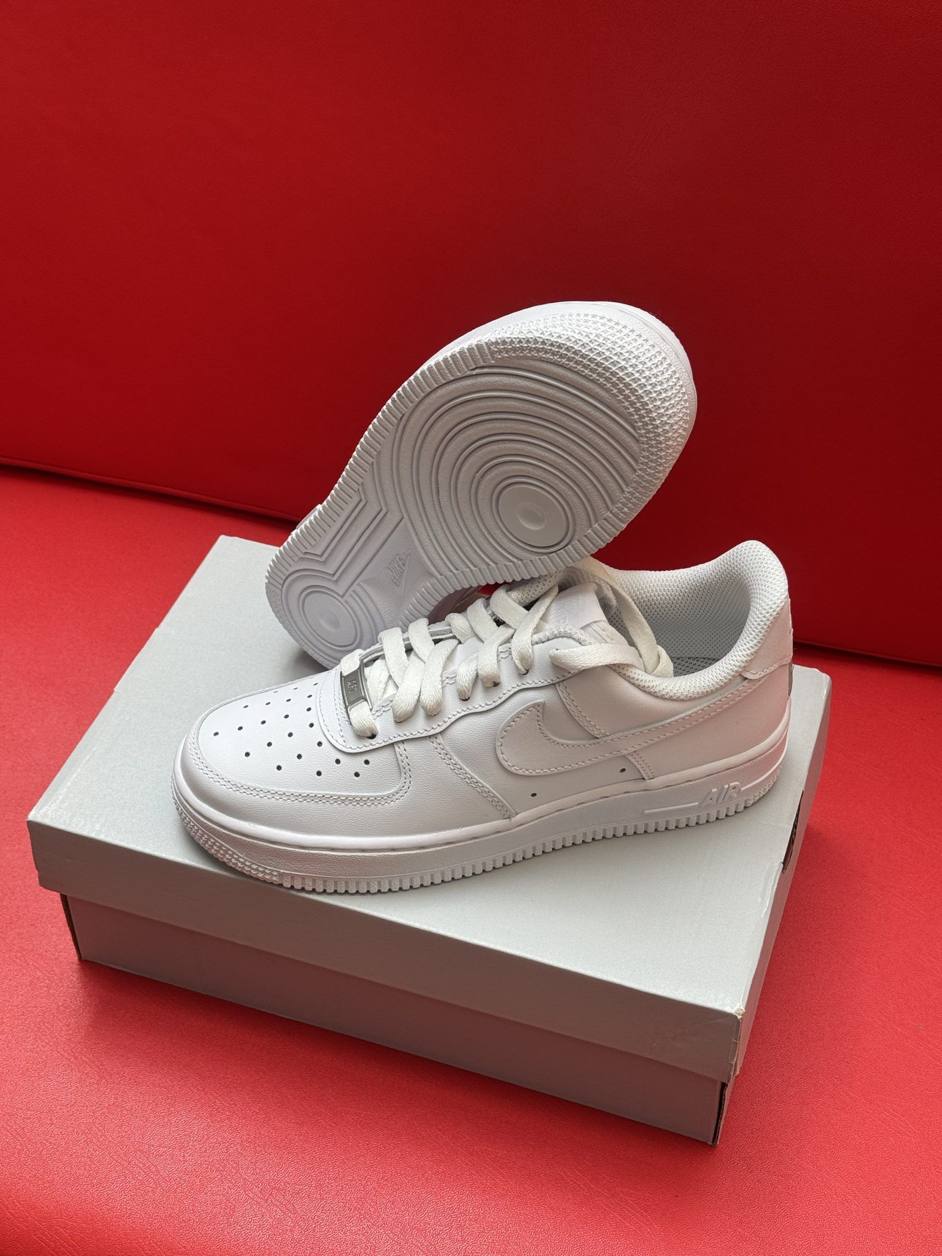 White Nike Air Force 1 sneakers, one upside down to show the sole, on a white box against a red background.