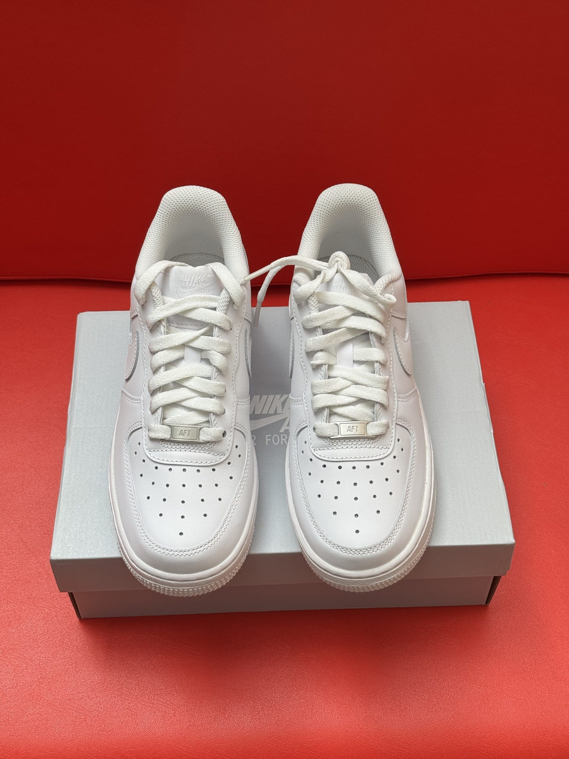 White Nike Air Force 1 sneakers on a white box, with the sole visible. Set against a red background.