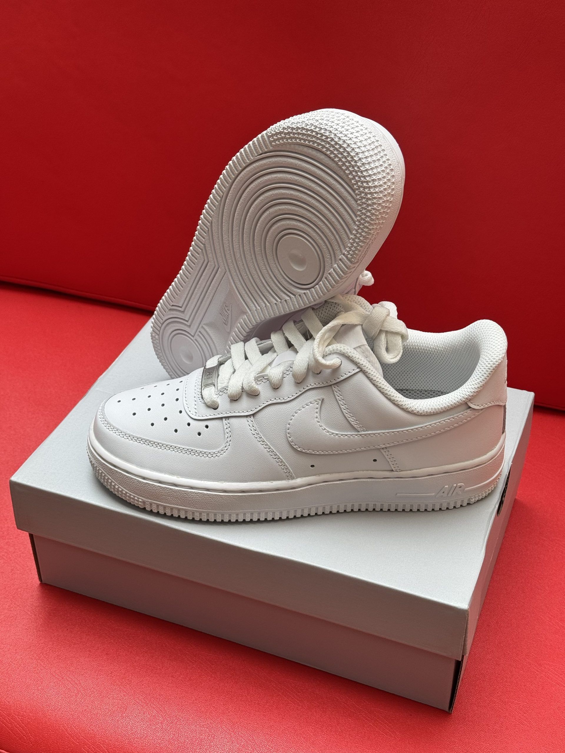 White Nike Air Force 1 sneakers on a white box, with the sole visible. Set against a red background.