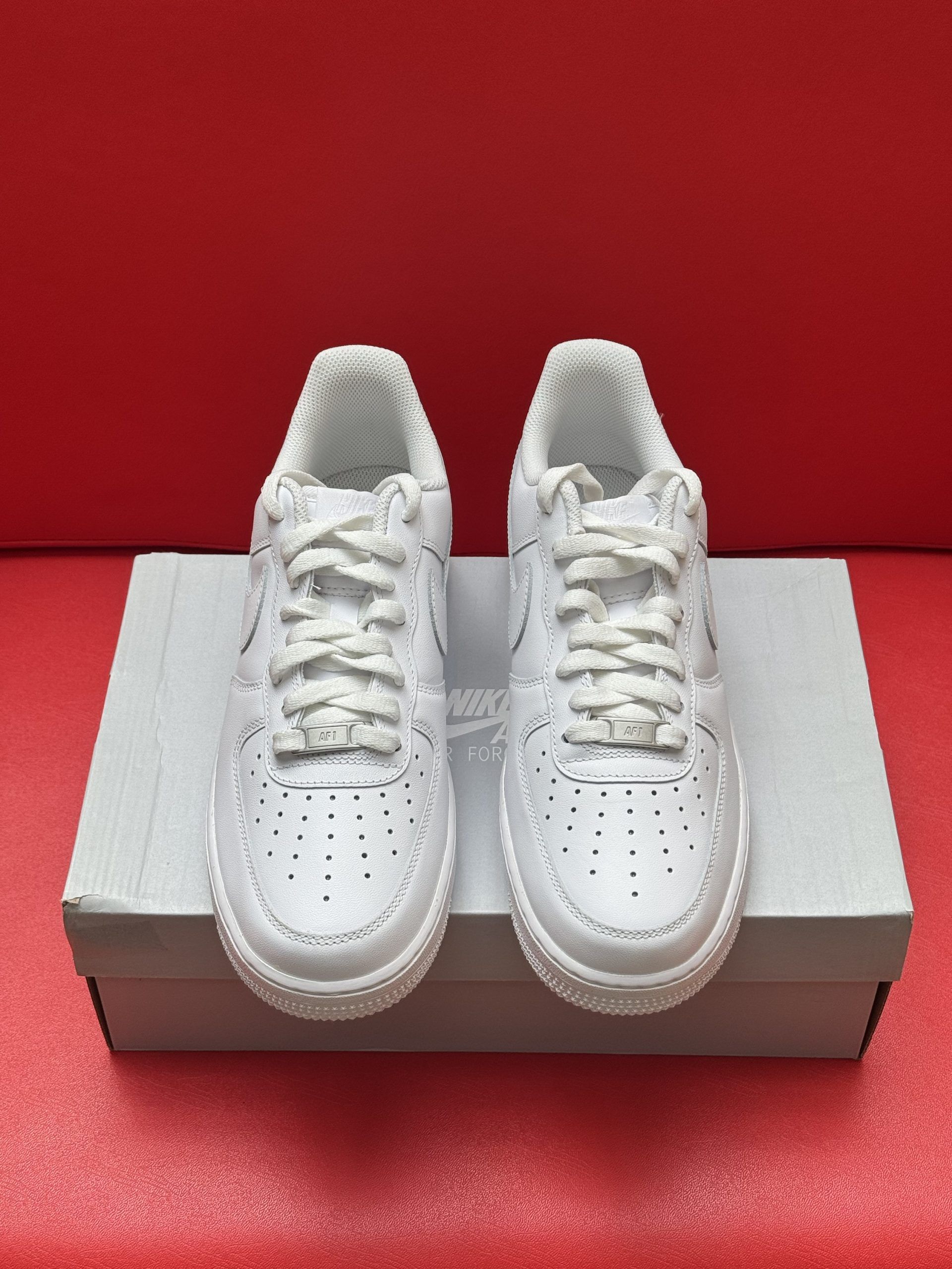 White Nike Air Force 1 sneakers on a box, sole up, against a red backdrop.