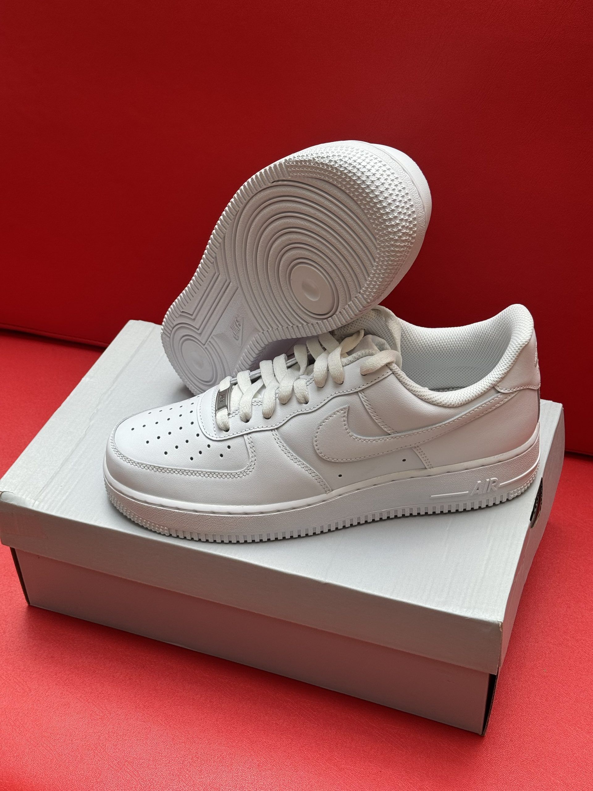 White Nike Air Force 1 sneakers on a box, sole up, against a red backdrop.
