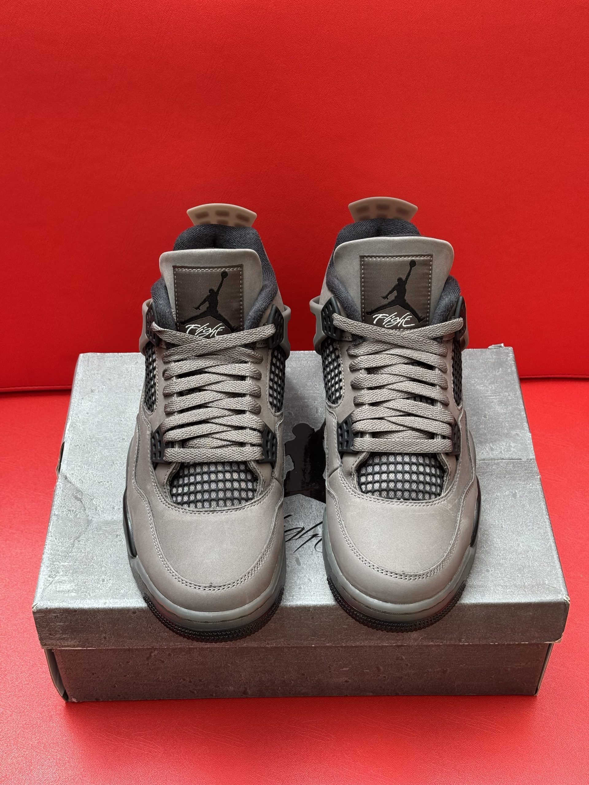 Dark gray Air Jordan 4 sneakers on a gray box, with the sole visible. Red backdrop.