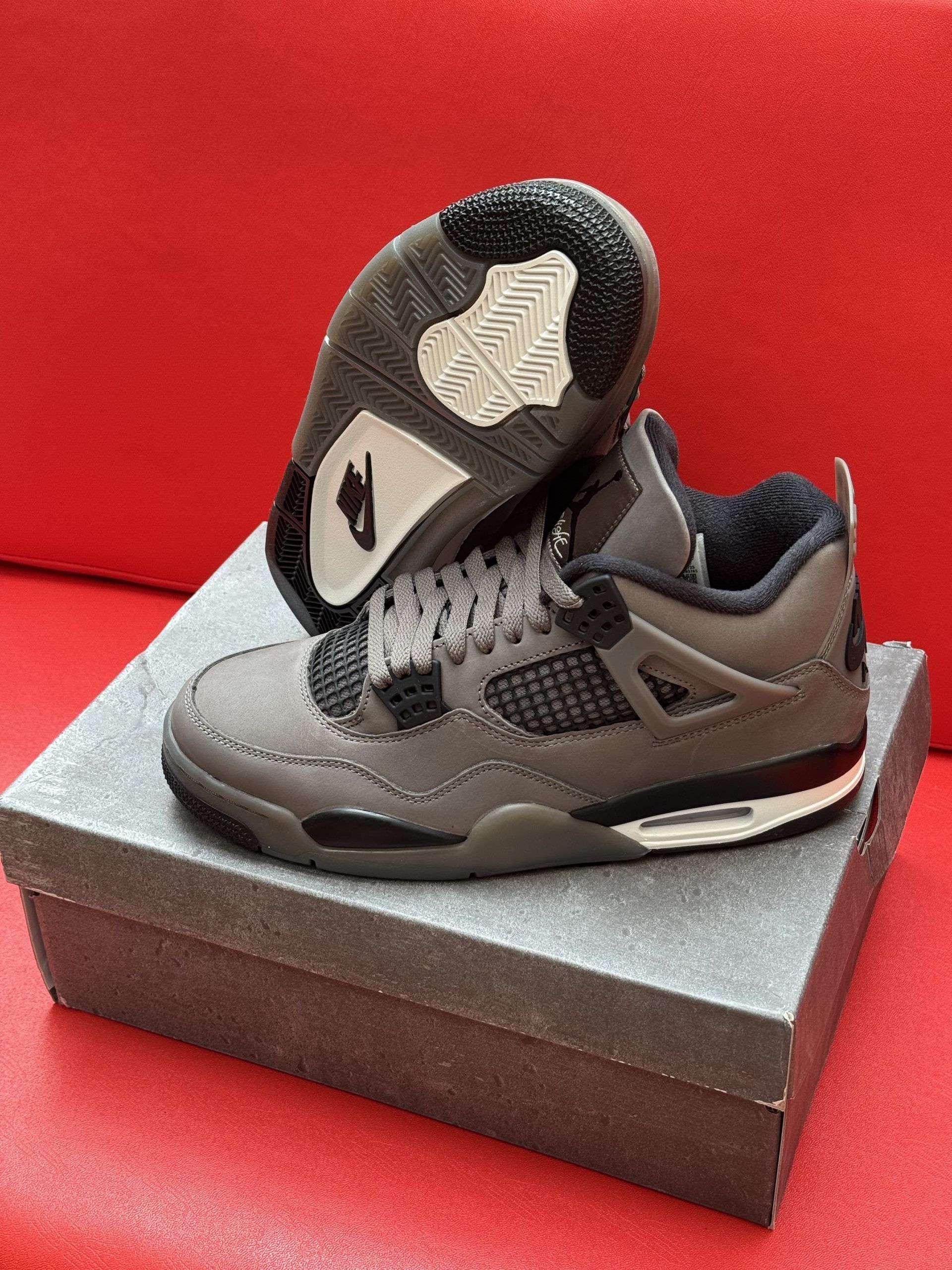 Dark gray Air Jordan 4 sneakers on a gray box, with the sole visible. Red backdrop.
