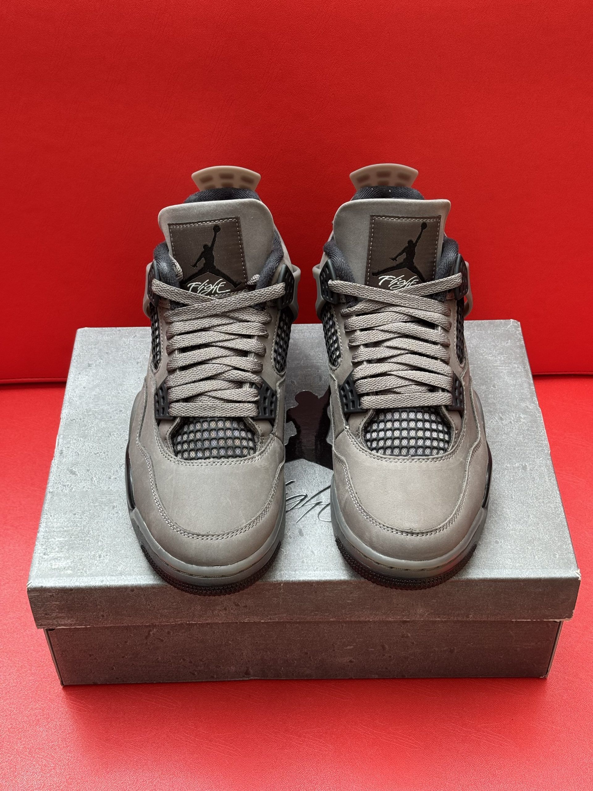 Pair of grey and black toddler Air Jordan 4 sneakers on a grey box, set against a red background.