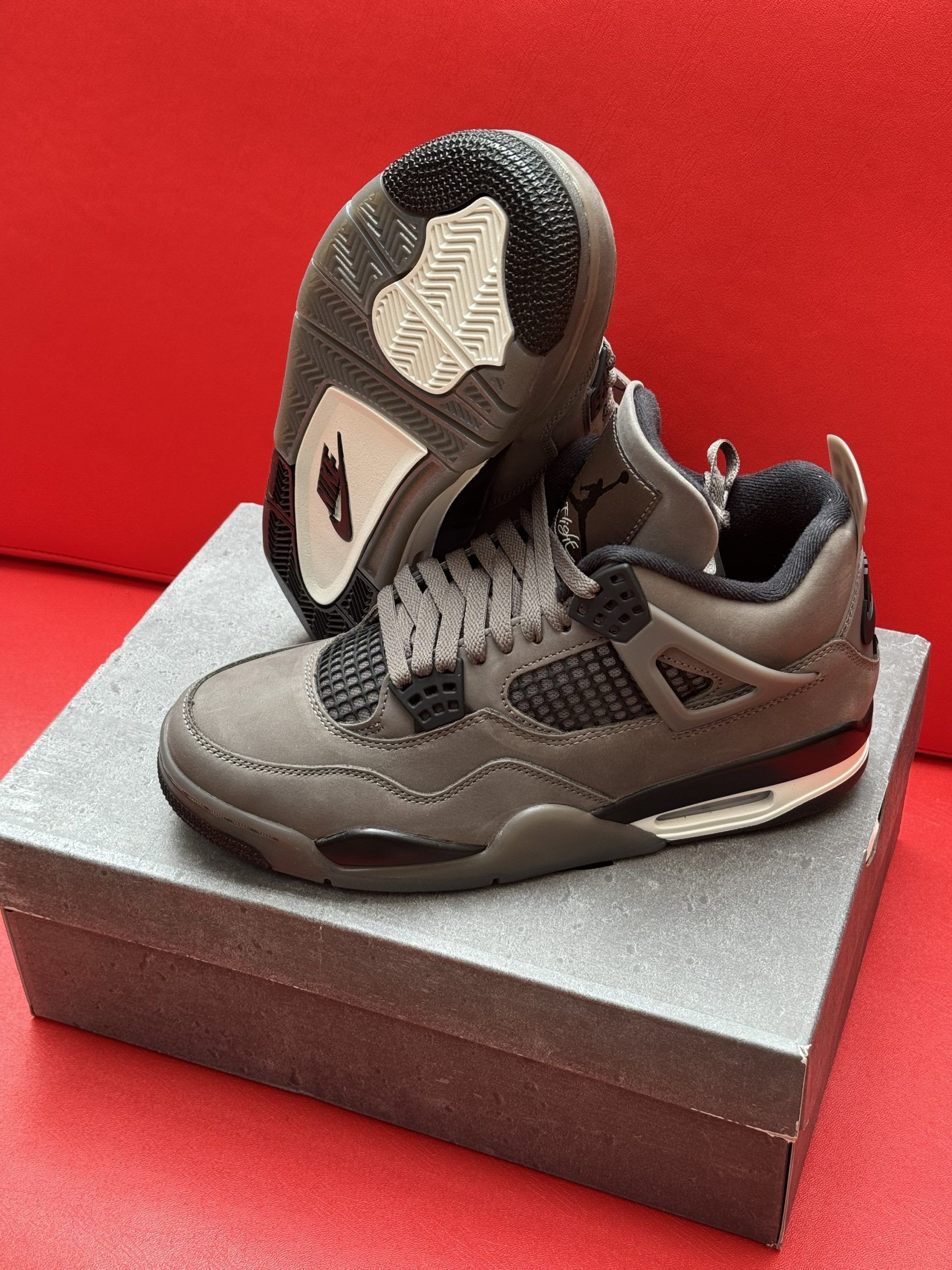 Pair of grey and black toddler Air Jordan 4 sneakers on a grey box, set against a red background.