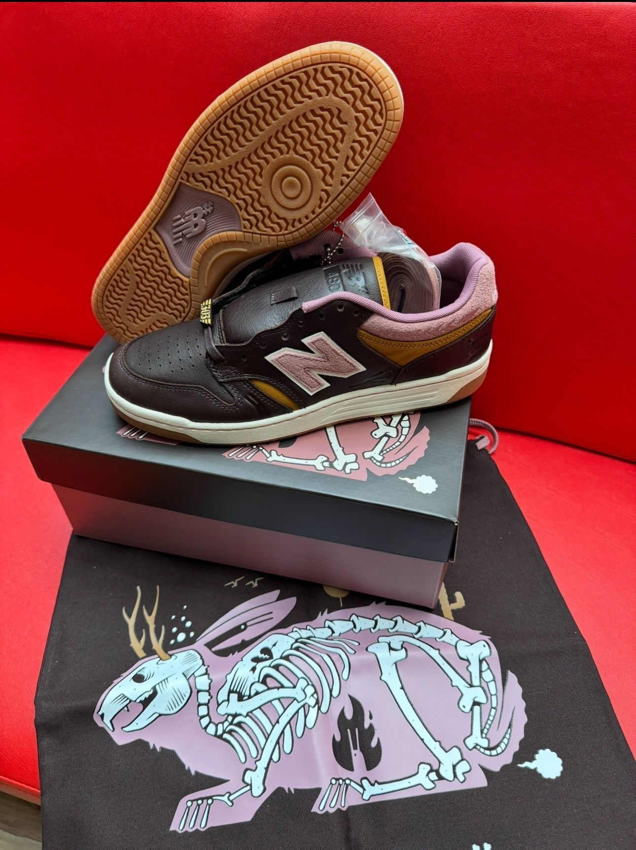 Brown New Balance sneakers with a bunny graphic on the box and tote bag, sitting on a red surface.