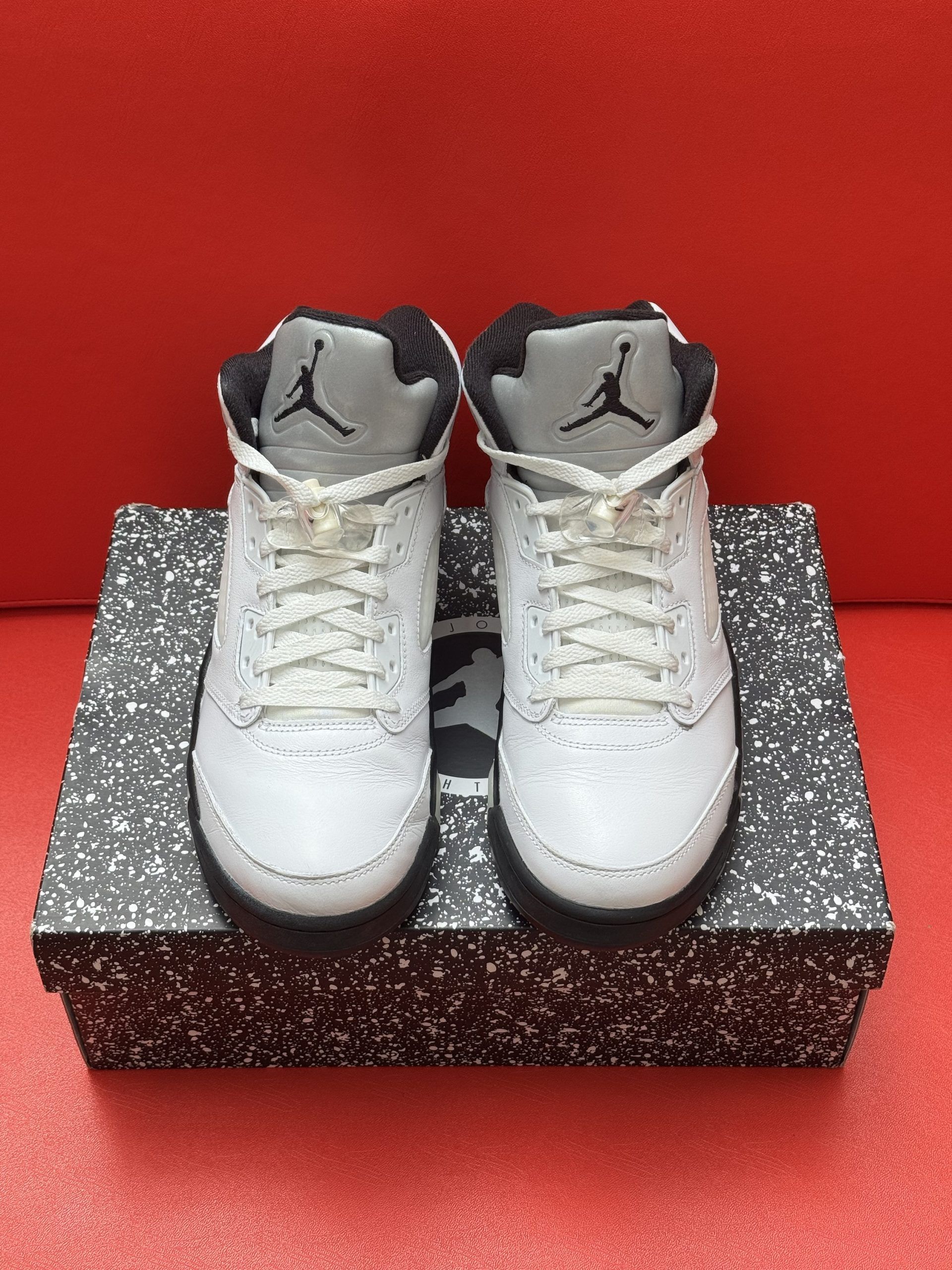 White and black Air Jordan 5 sneakers on speckled box, sole in the background, set on a red surface.