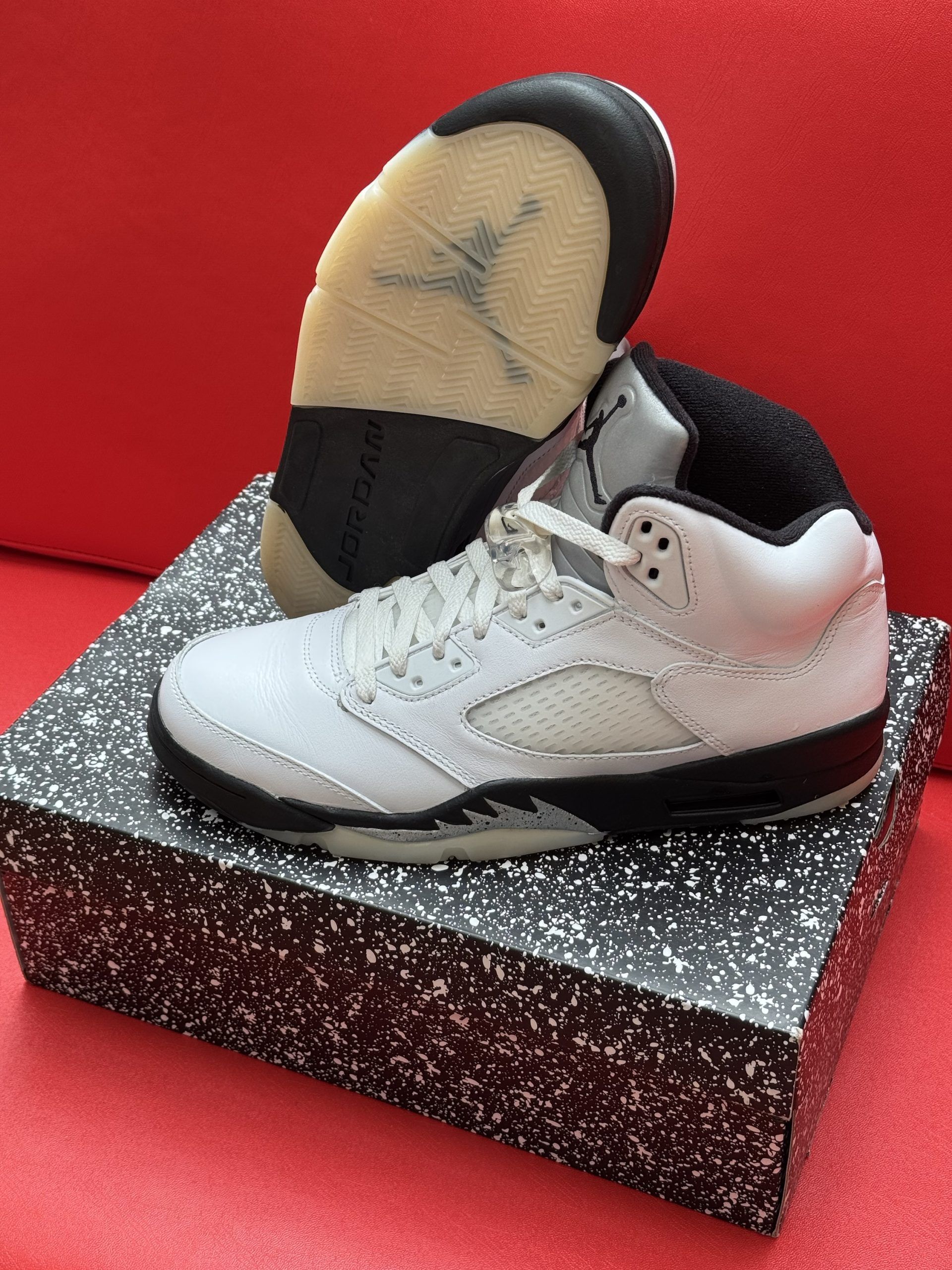 White and black Air Jordan 5 sneakers on speckled box, sole in the background, set on a red surface.