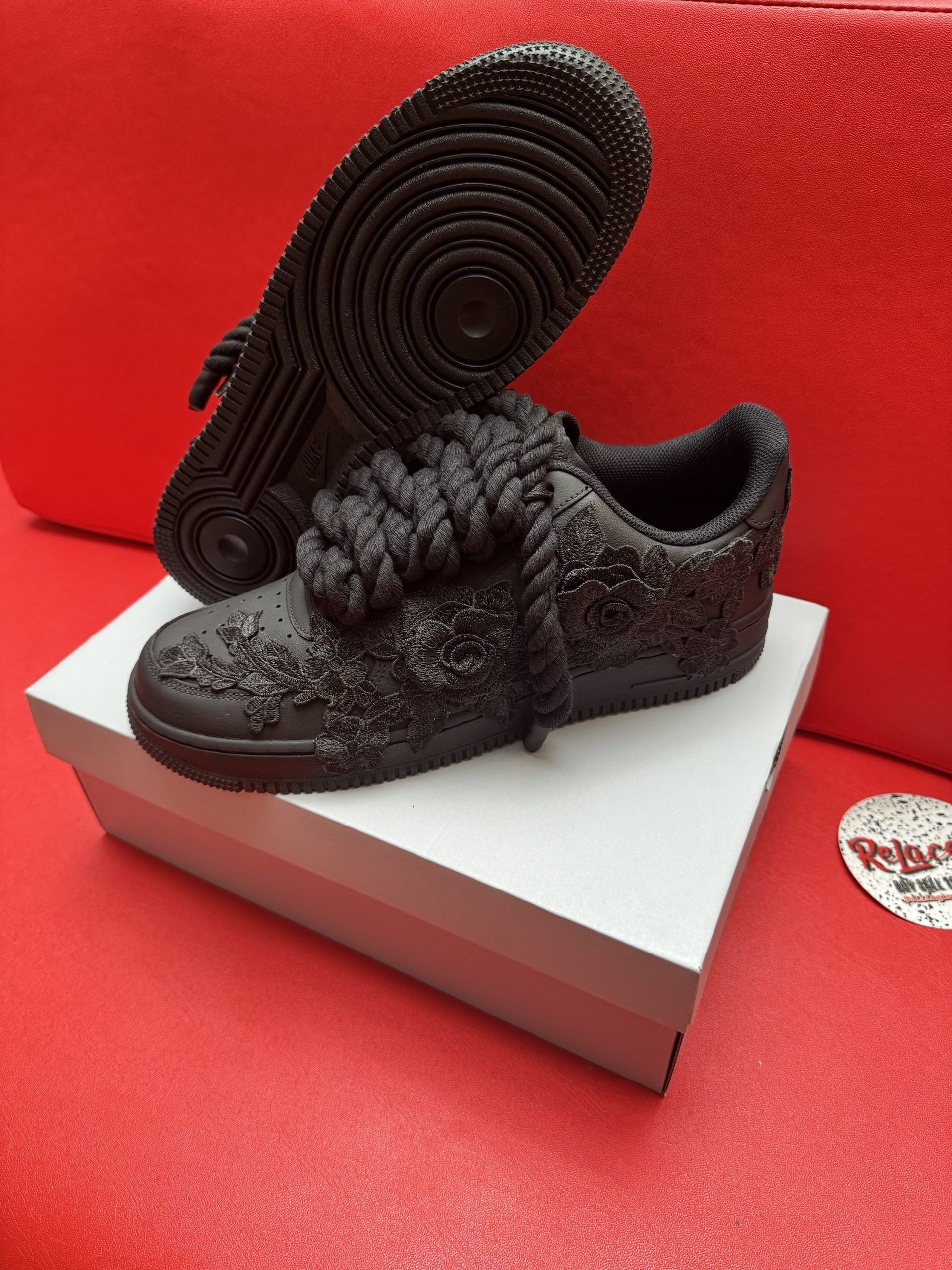 Black textured sneakers on white box, red background. Sole of one shoe visible.